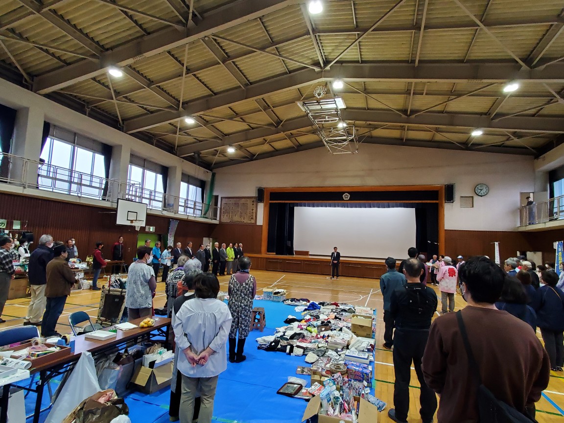 体育館にブルーシートが敷かれた上に様々な品物が並べられており、関係者がステージ前で話をする高橋市長見ている様子の写真