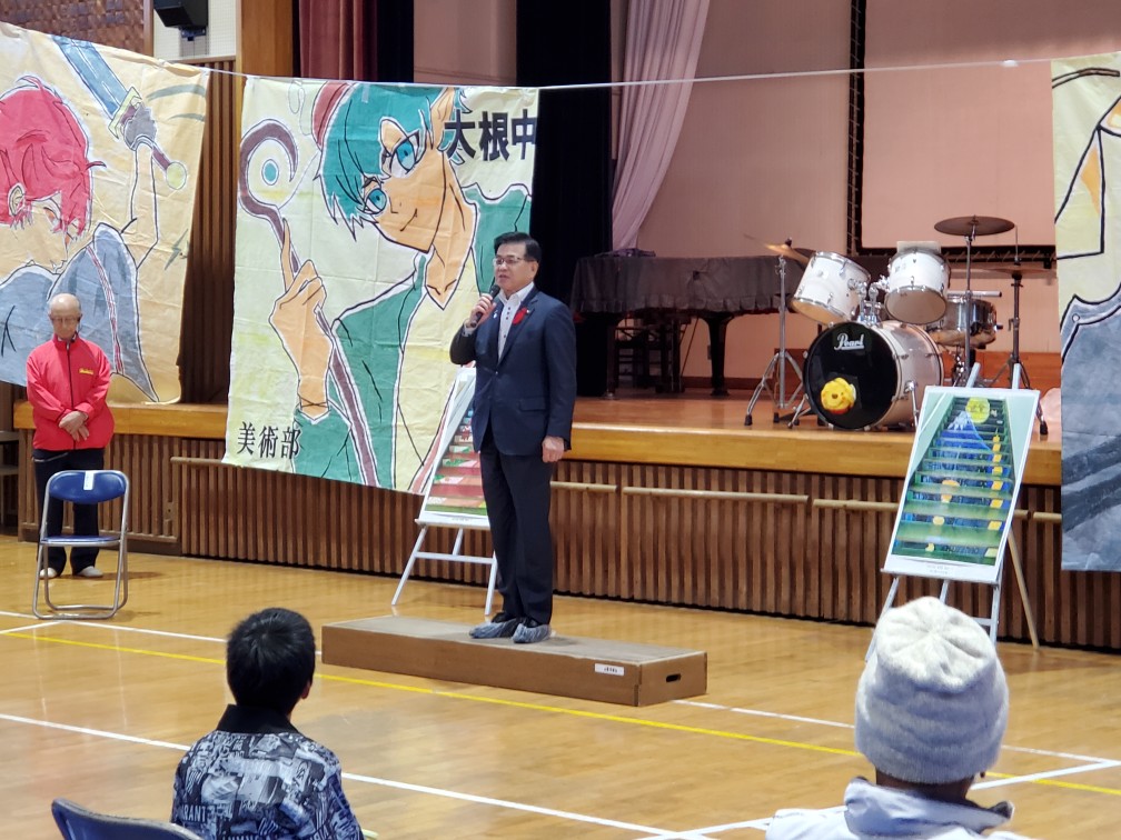 アニメ風のイラストが大きく描かれたものが飾られており、その前でマイクを持ってスピーチをする高橋市長の写真