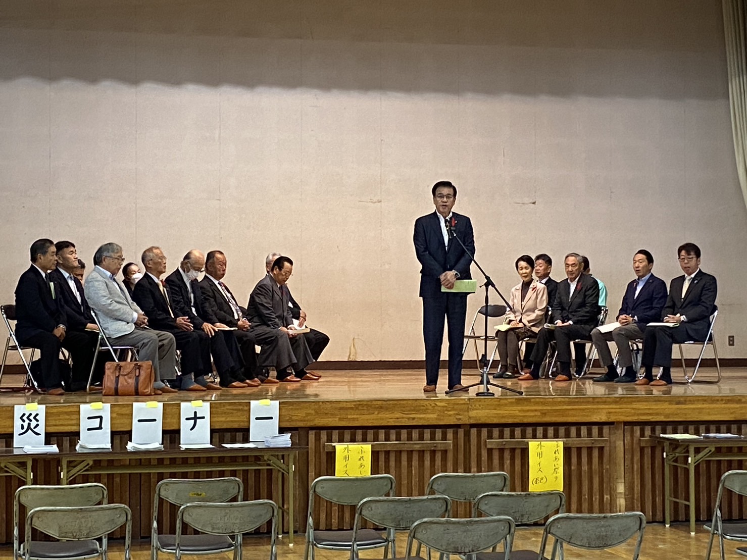 椅子が並べられたステージの上で、マイクに向かって話す高橋市長と、その両側に椅子に座る人々の写真