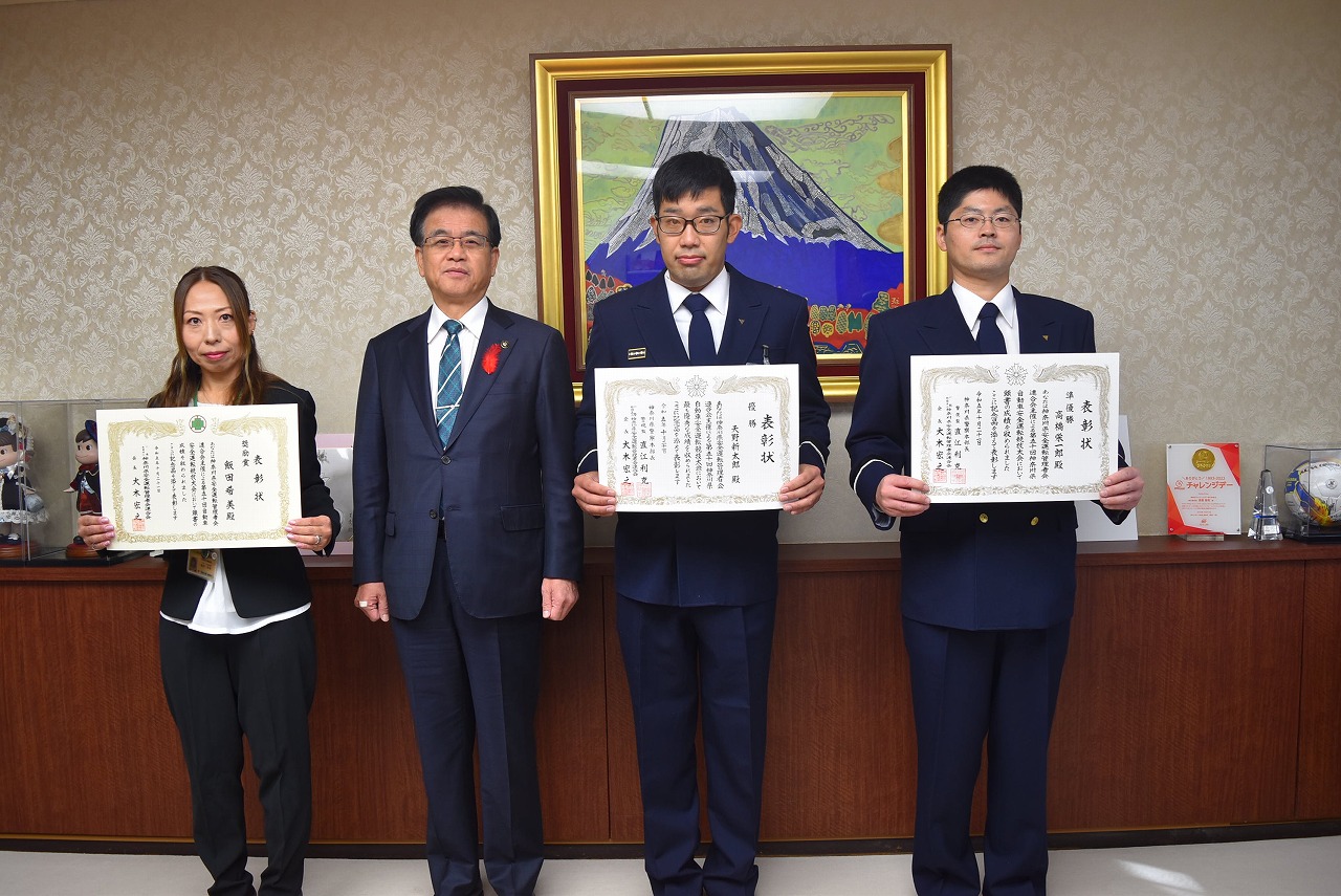 富士山の絵画を背景に、市長と並んで、神奈川県自動車安全運転競技大会で入賞した制服姿の男性2名とスーツ姿の女性が賞状を手に笑顔で写っている記念写真