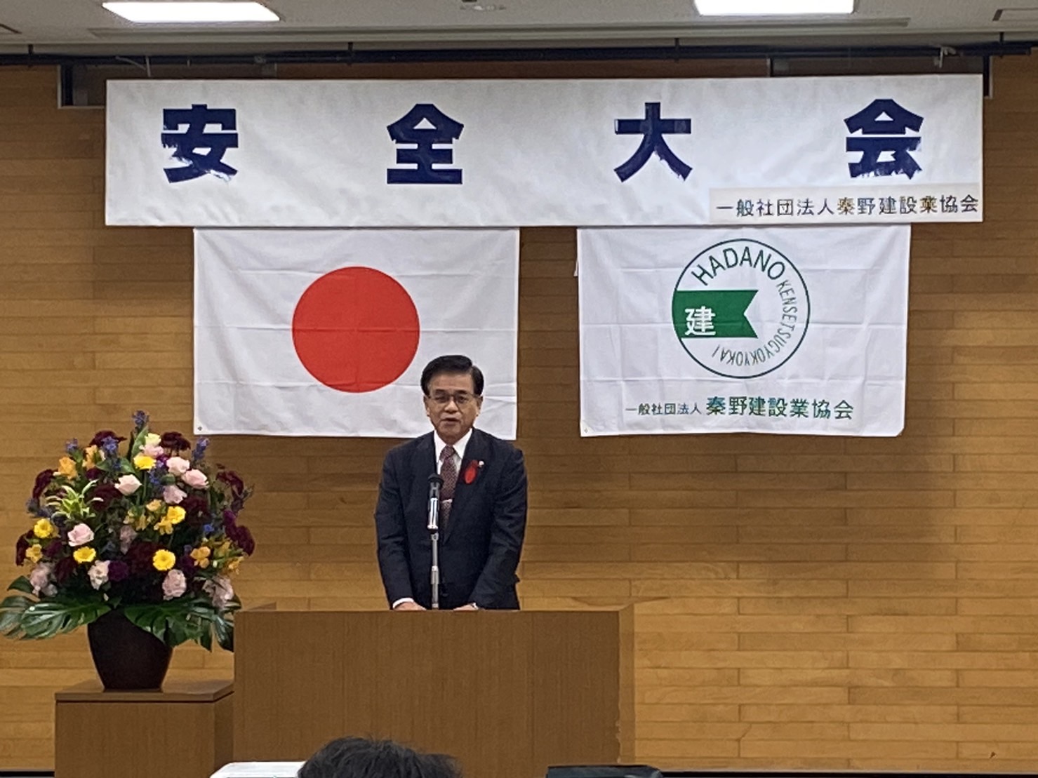 安全大会の壇上で、日の丸と建設業協会の旗を背に、スーツ姿の市長がマイクの前に立って挨拶をしている写真