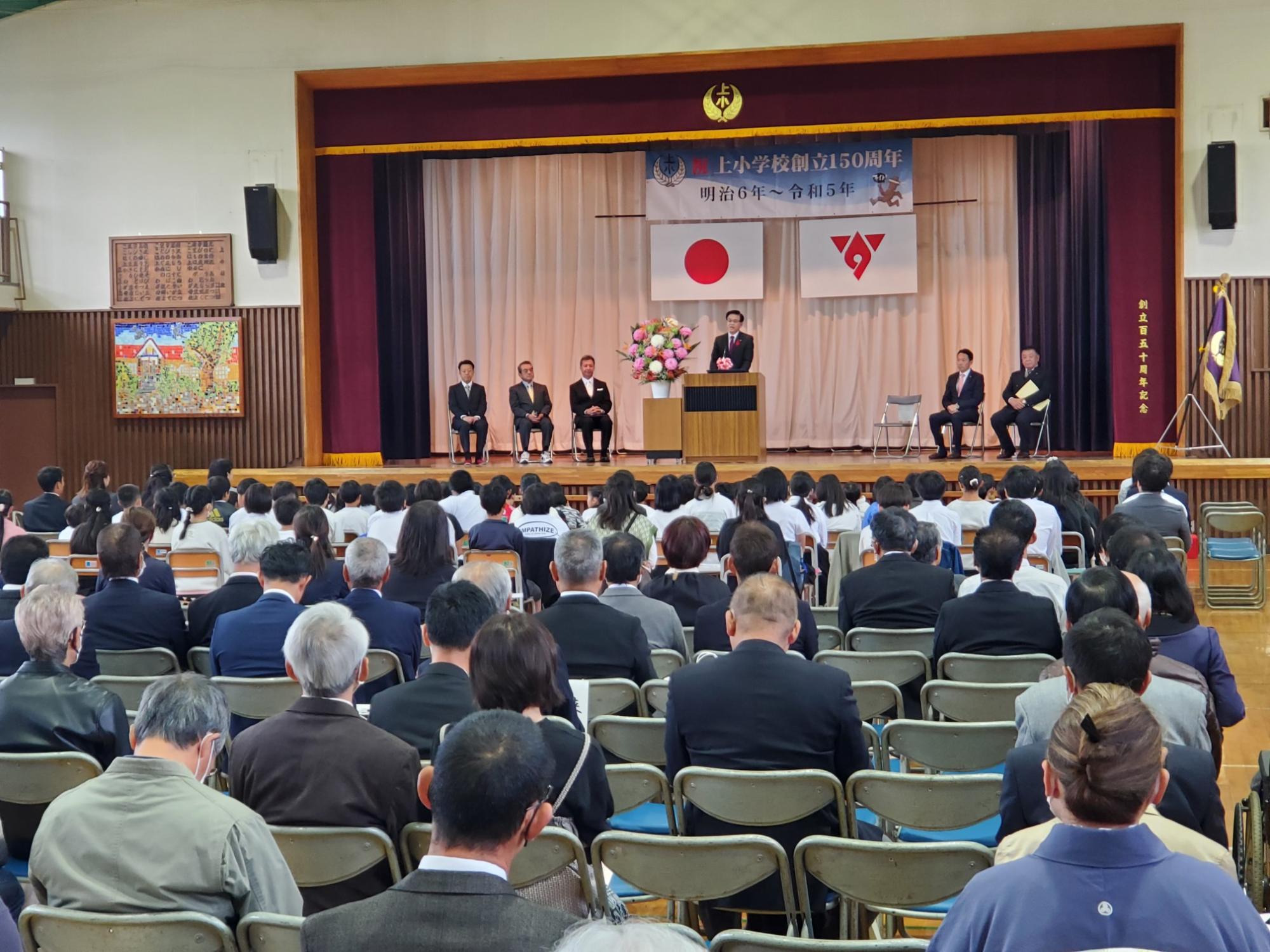 上小学校創立150周年記念式典の横断幕と日の丸、秦野市の市旗が掲示された舞台に関係者の方5名が椅子に座り、壇上で挨拶する市長と多くの来場者が客席に座って話を聞いている様子を後方から写した写真