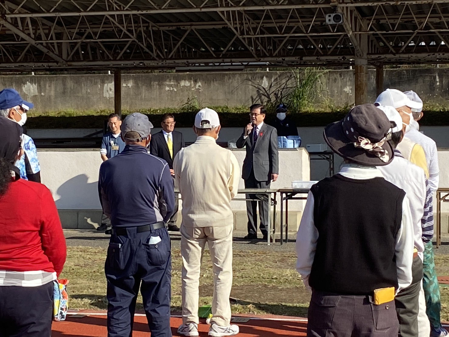 晴天の下、屋外の会場でカジュアルな服装の参加者の方々が整列する中、市長がマイクを手に挨拶を行っている様子を写したターゲット・バードゴルフ協会市長杯の写真