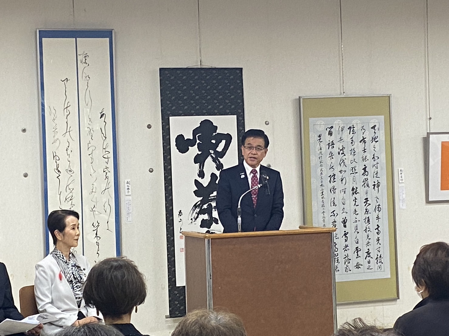 書道の作品の展示を背景に、市長が壇上で挨拶を行っている様子の写真