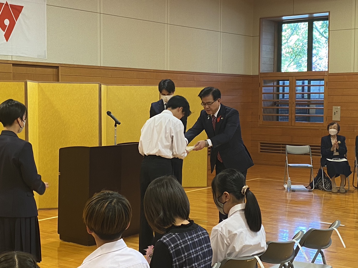 黄色いパネルを背景にした式典会場で、受賞者の男子生徒が市長から表彰を受けている様子を写した写真