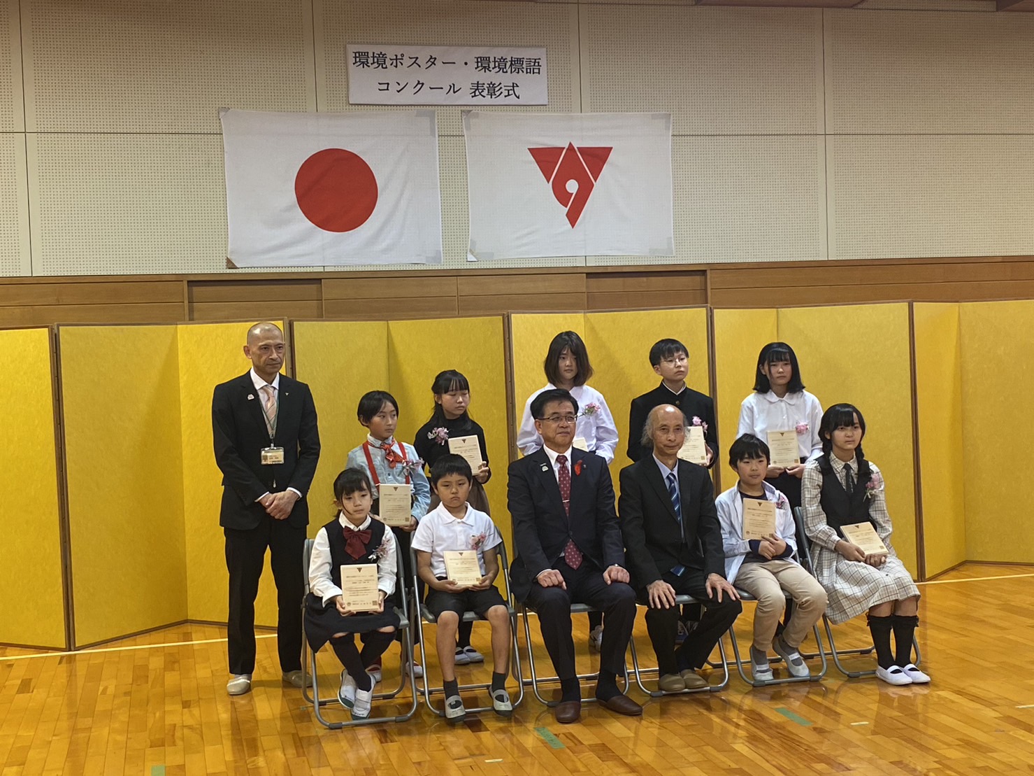 「環境ポスター・環境標語コンクール表彰式」の掲示と日本国旗と秦野市の市旗が掲示され、黄色いパネルを背景に、賞状を持った子どもたちと市長、関係者の方々が2列に並び写っている記念写真