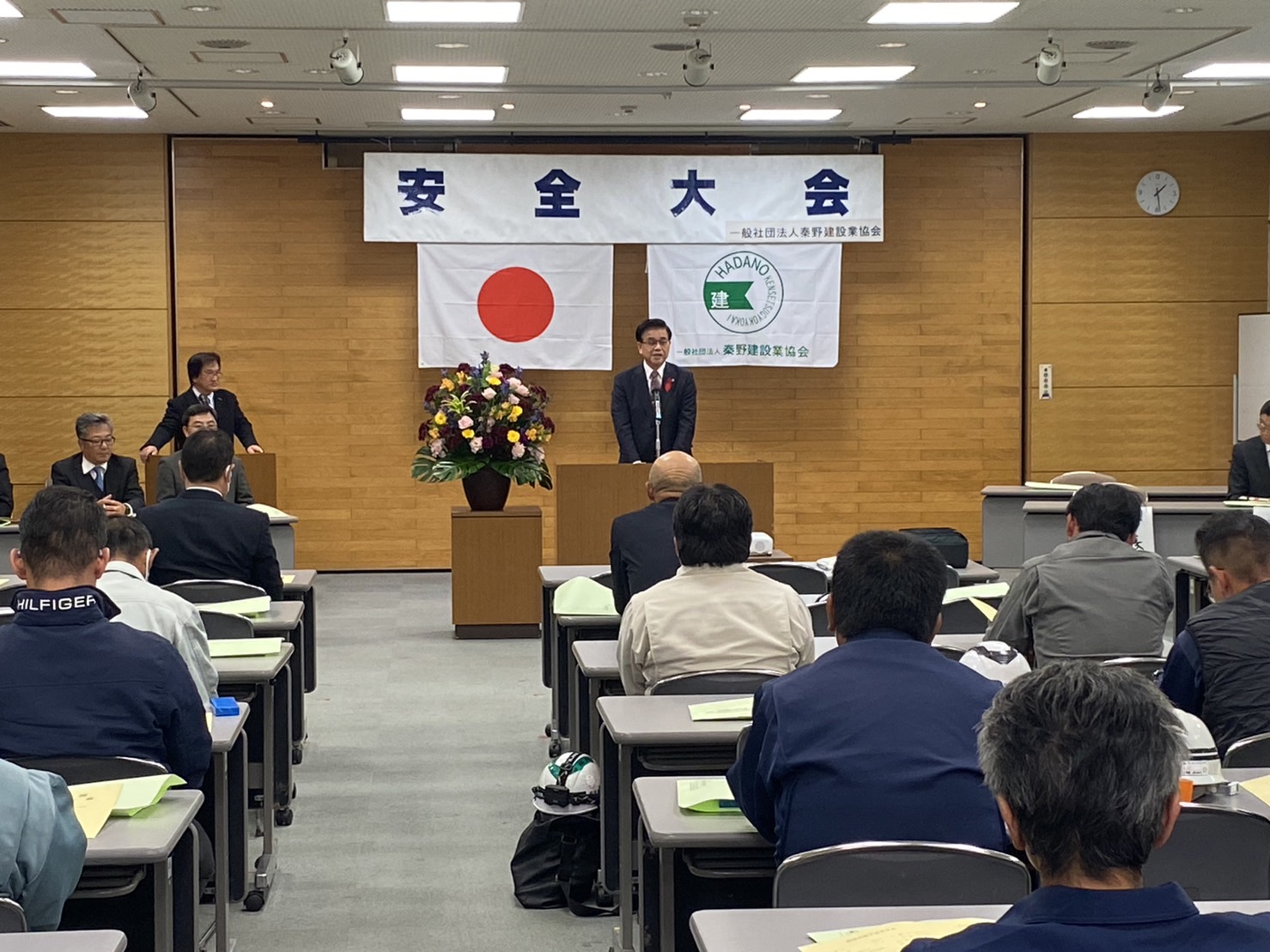 安全大会の壇上で、日の丸と建設業協会の旗を背に、スーツ姿の市長がマイクの前に立って挨拶をしており、会場には多くの参加者が席に着き、話を聞いている建設業安全大会の会場の様子を後方から写した写真