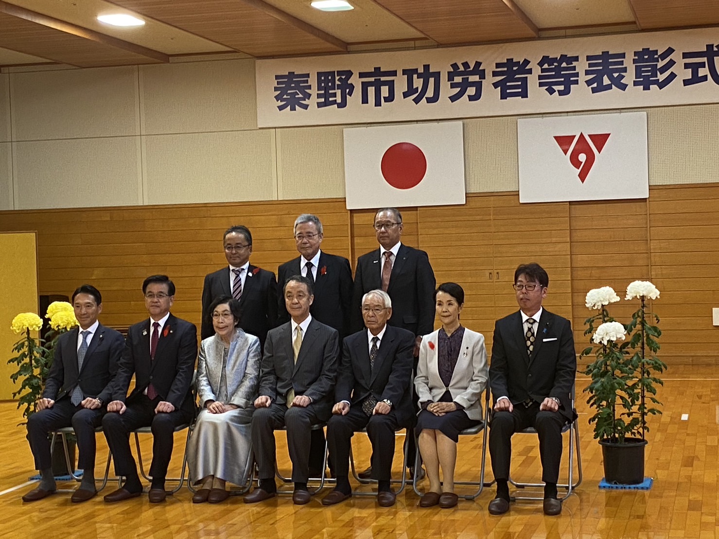 日本の国旗が掲げられた室内で、「秦野市功労者等表彰式」と書かれた横断幕の下でスーツや和服を着用した市長と男女が並んで記念写真