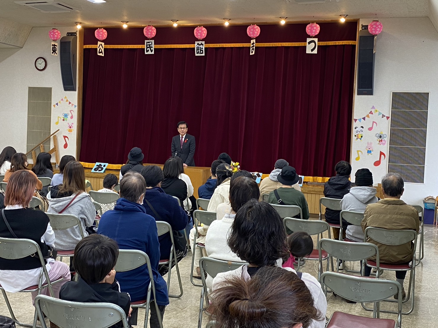 天井に「東公民館まつり」と一文字ずつ飾りと共に吊り下げられた舞台の前で、市長が壇上に立って挨拶を行っており、会場の観客席に多くの来場者が席について話を聞いている様子を後方から写した写真