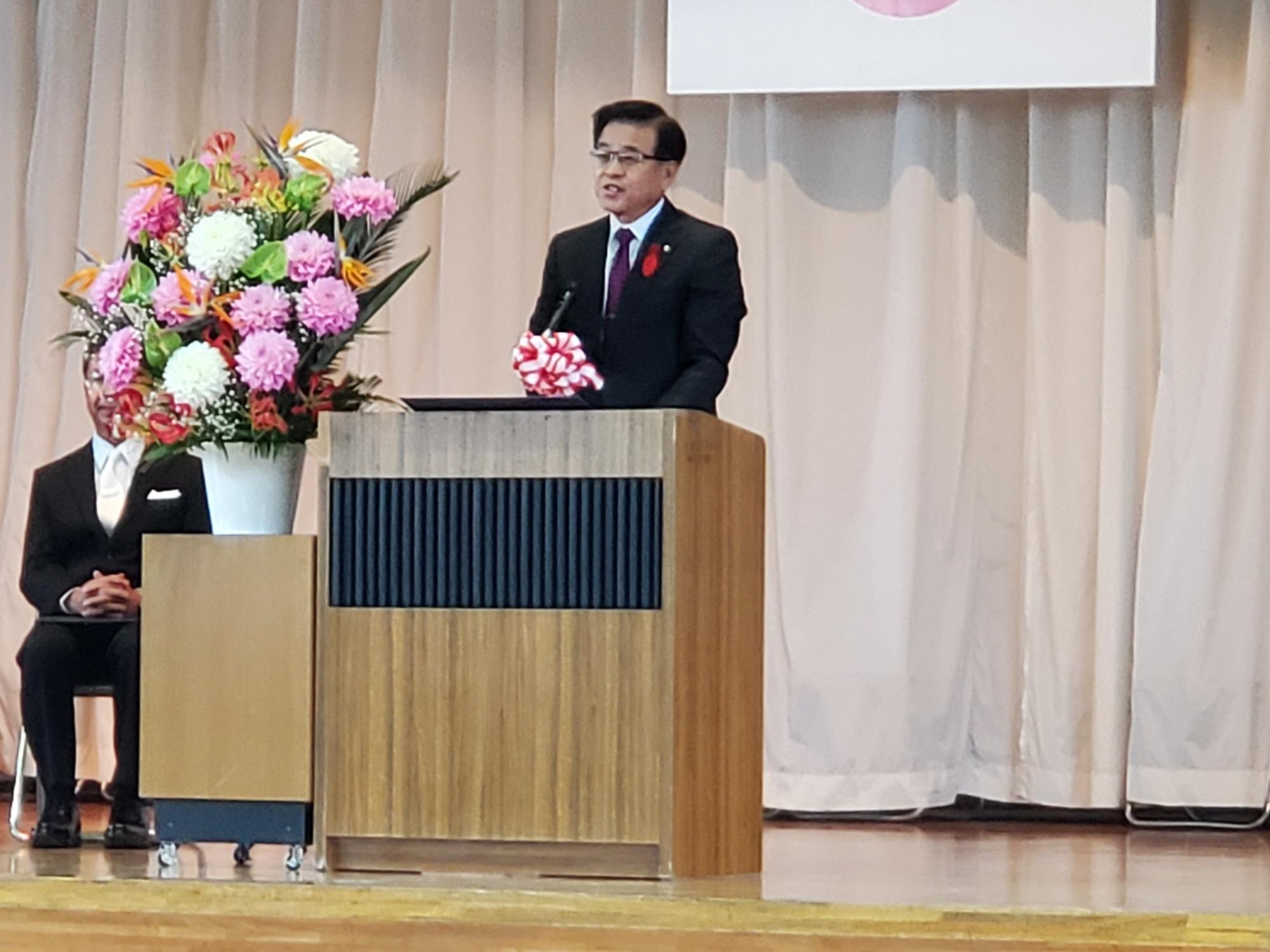 華やかな生花が飾られた壇上で、市長が挨拶を行っている様子の写真