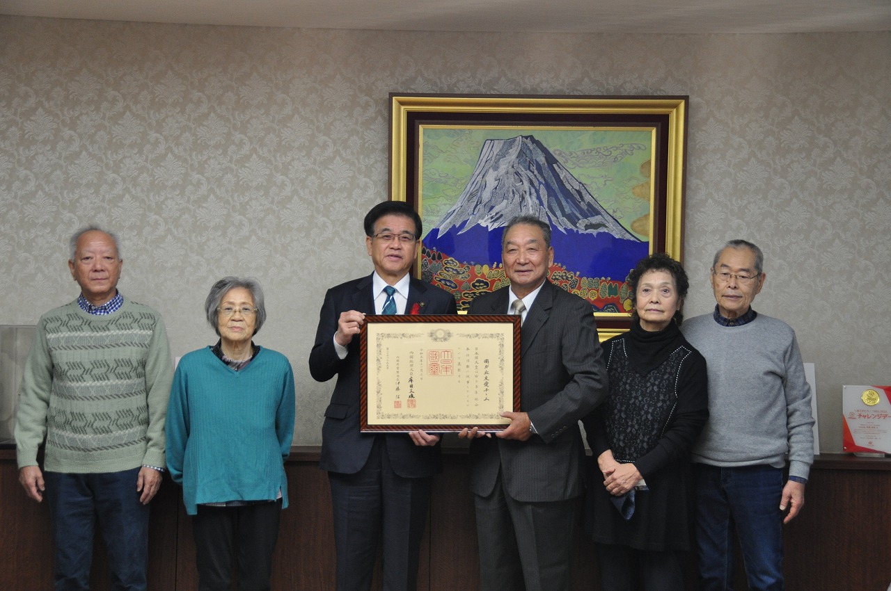 富士山の絵画を背景に、中央に立つ市長とスーツ姿の男性が並んで褒章の賞状を持ち、関係者の方々と笑顔で写っている記念写真