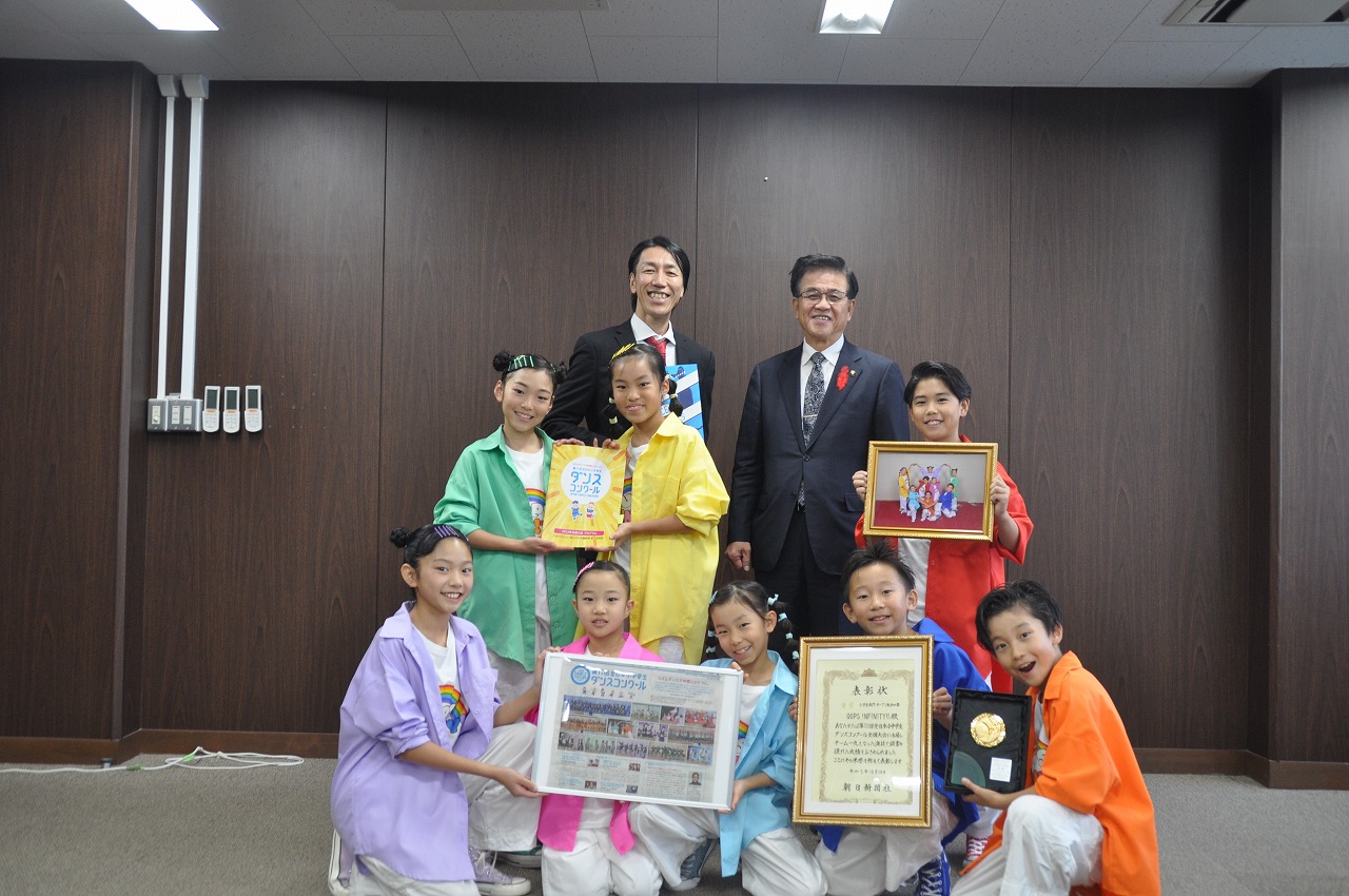 賞状や盾、写真などを手にしたカラフルなダンスの衣装を身に着けたMK☆ダンスの子ども達が市長と関係者の男性と並んで笑顔で写っている記念写真