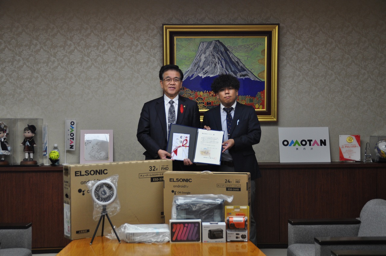 富士山の絵画を背景に、寄贈された電子機器等の商品が机の上に並べられ、市長と久宗店長が寄贈を示す証書を持ち笑顔で写っている記念写真