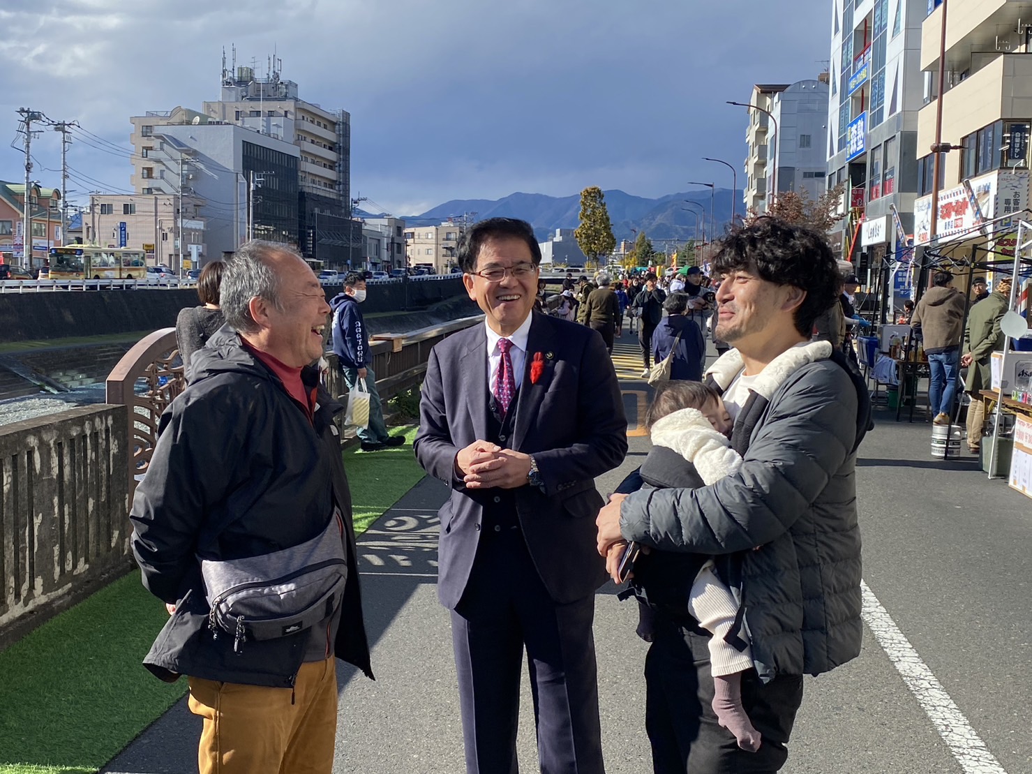 川沿いの道路で、市長が年配の男性と幼い子どもを抱いた男性と笑顔で交流している様子の写真