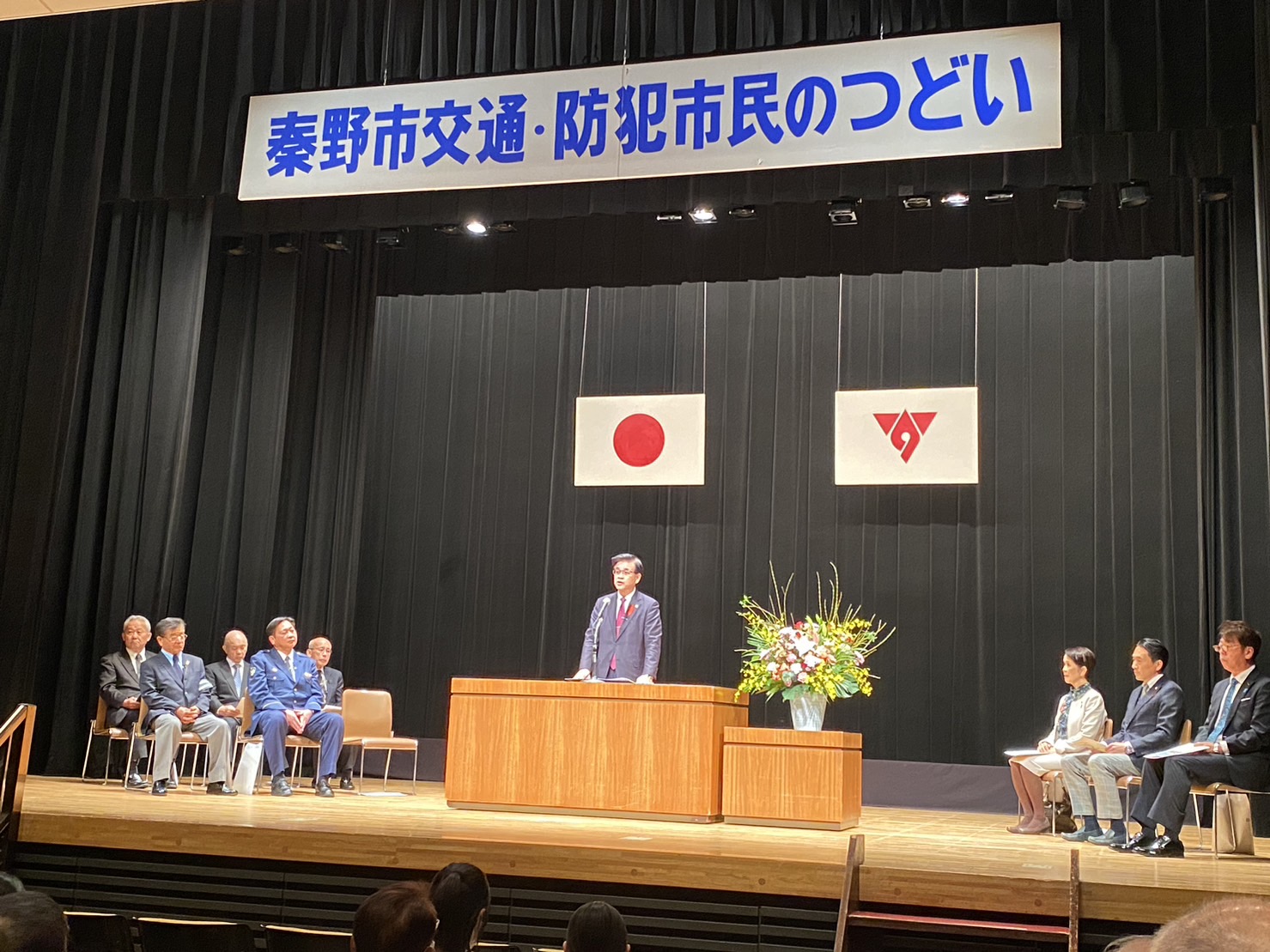 「秦野市交通・防犯市民のつどい」の横断幕と日本国旗と秦野市の市章が掲げられた会場で、左右にスーツ姿の関係者が着席し、市長が演壇の前に立ち話をしている様子の写真