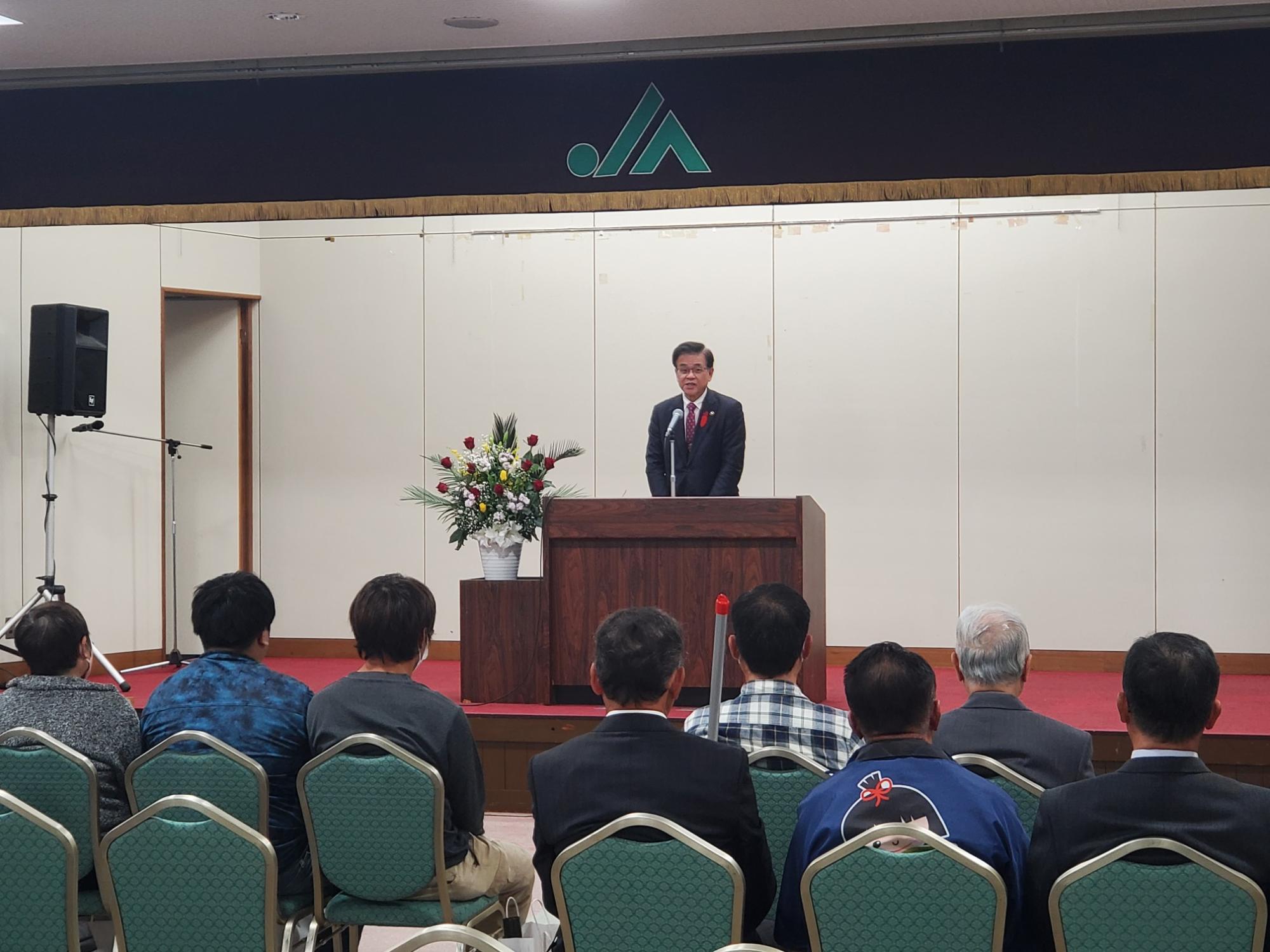 秦野市農産物品評会ほう賞授与式の会場で壇上のマイク前に市長が立ち話をしており、参列者たちが着席して話を聞いている様子の写真