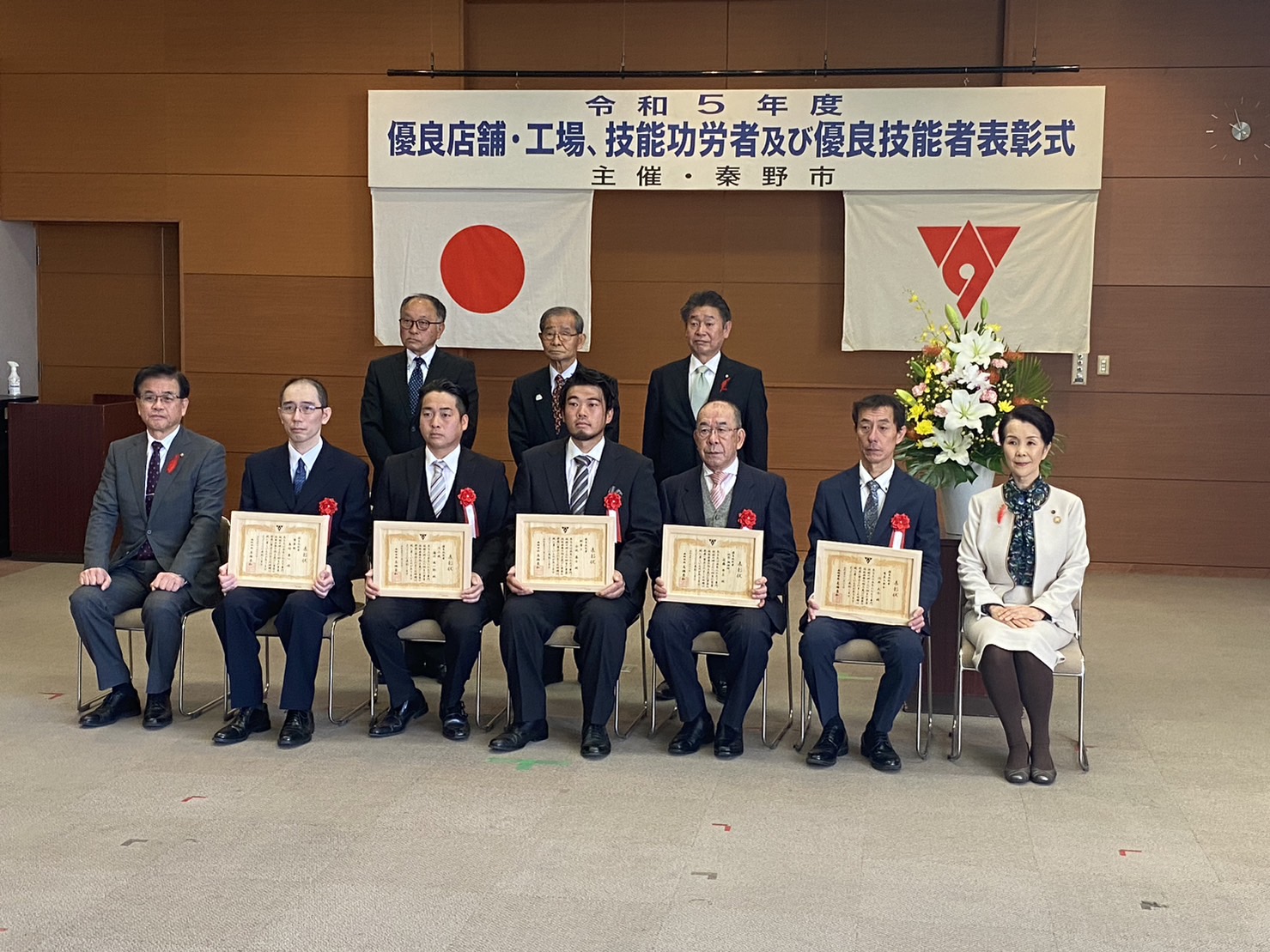 『令和5年度 優良店舗・工場、技能功労者及び優良技能者表彰式』の横断幕の前で、受賞者5名が表彰状を手に持ち、関係者とともに記念撮影を行っている様子の写真