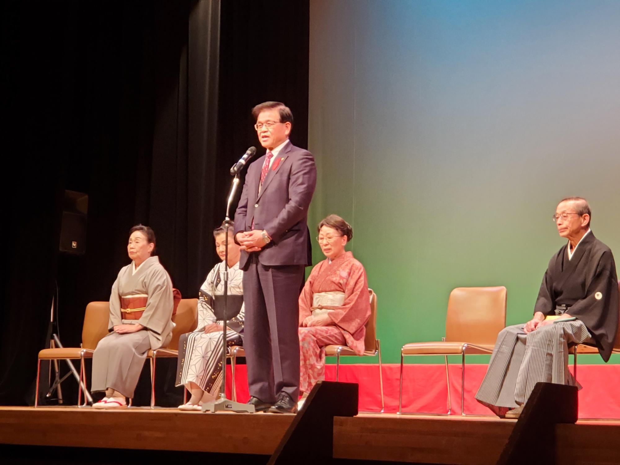 舞台上でスーツ姿の市長がマイクを前に話し、後方には和装の女性3名と袴姿の男性1名が椅子に座っている民謡民舞大会の様子の写真