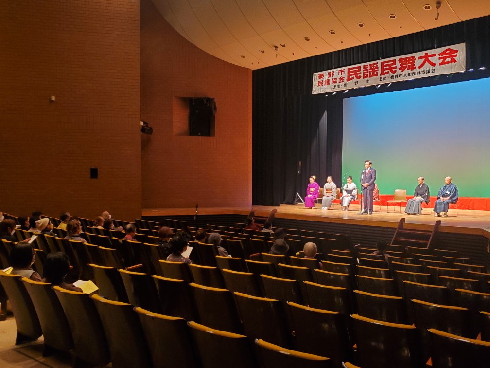 民謡民舞大会と書かれた横断幕が掲げられ、舞台後方には和装の女性4名と袴姿の男性2名が椅子に座っており、舞台中央で、市長がマイクの前に立ち話をしており、観客席には多くの人が座っている民謡民舞大会会場の様子を写した写真