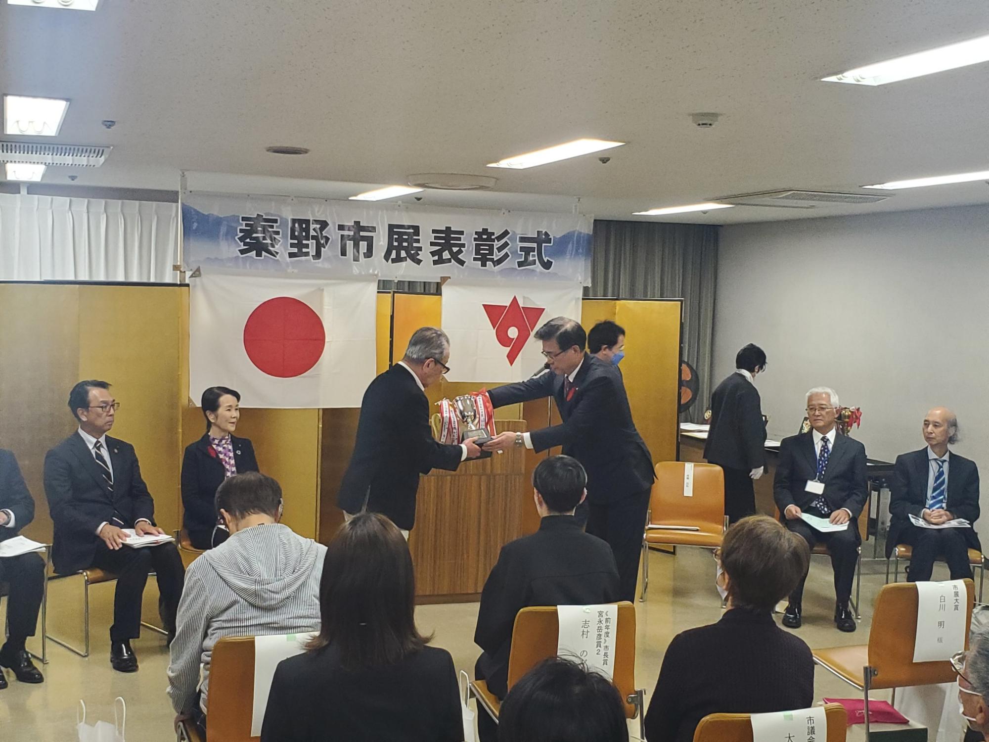 「秦野市展表彰式」の横断幕と日本国旗と秦野市の市章が掲げられた会場で、市長が受賞者にトロフィーを手渡している様子の写真