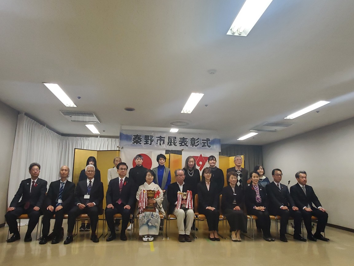 「秦野市展表彰式」の横断幕と日本国旗と秦野市の市章が掲げられた金屏風の前で、トロフィーを持った受賞者と関係者が2列に並び、で記念撮影を行っている様子の写真