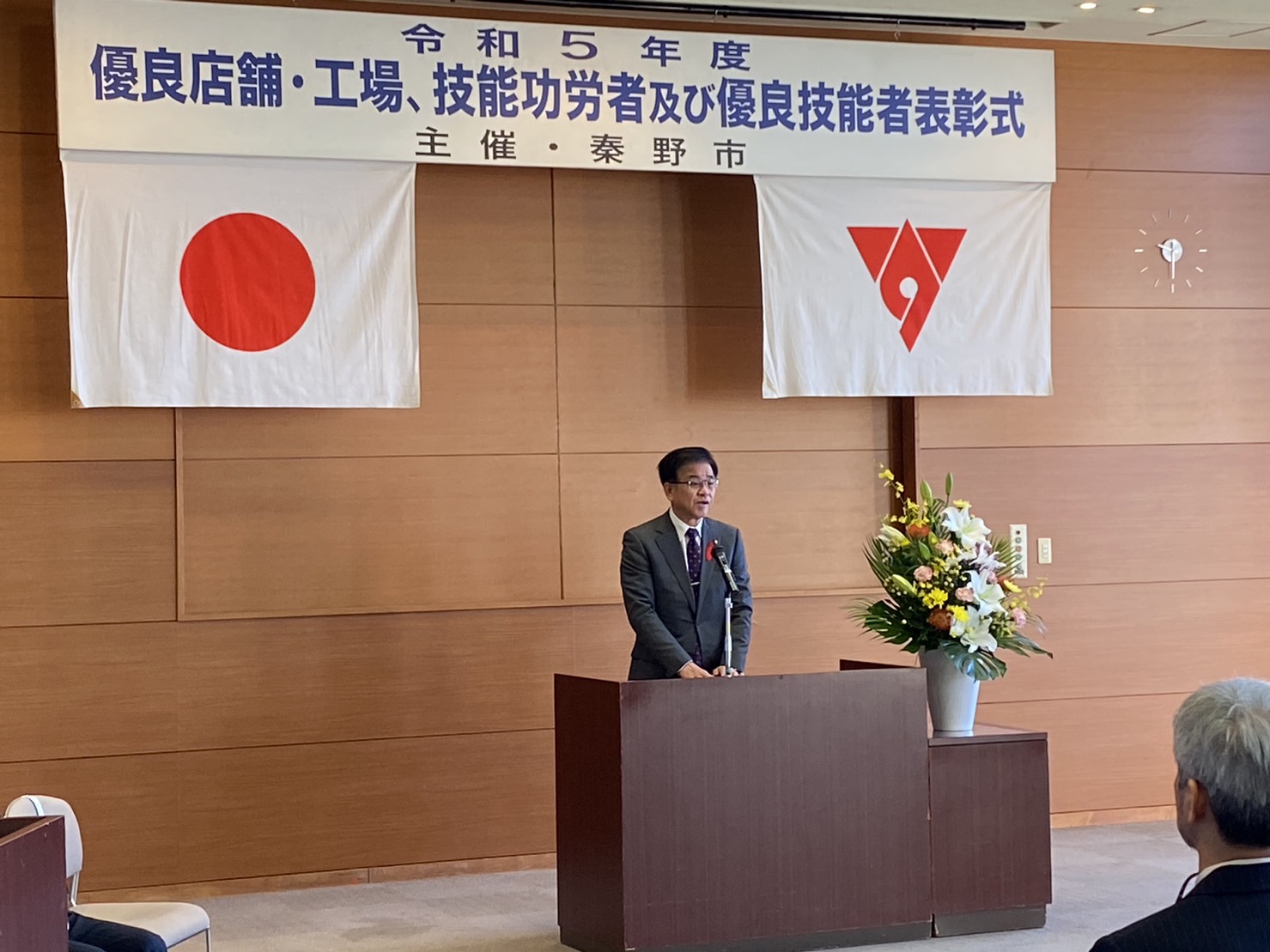 『令和5年度 優良店舗・工場、技能功労及び優良技能者表彰式』と書かれた横断幕が掲げられ、日本国旗と秦野市の市章が掲げられ、壇上に立つ市長が話をしている様子の写真