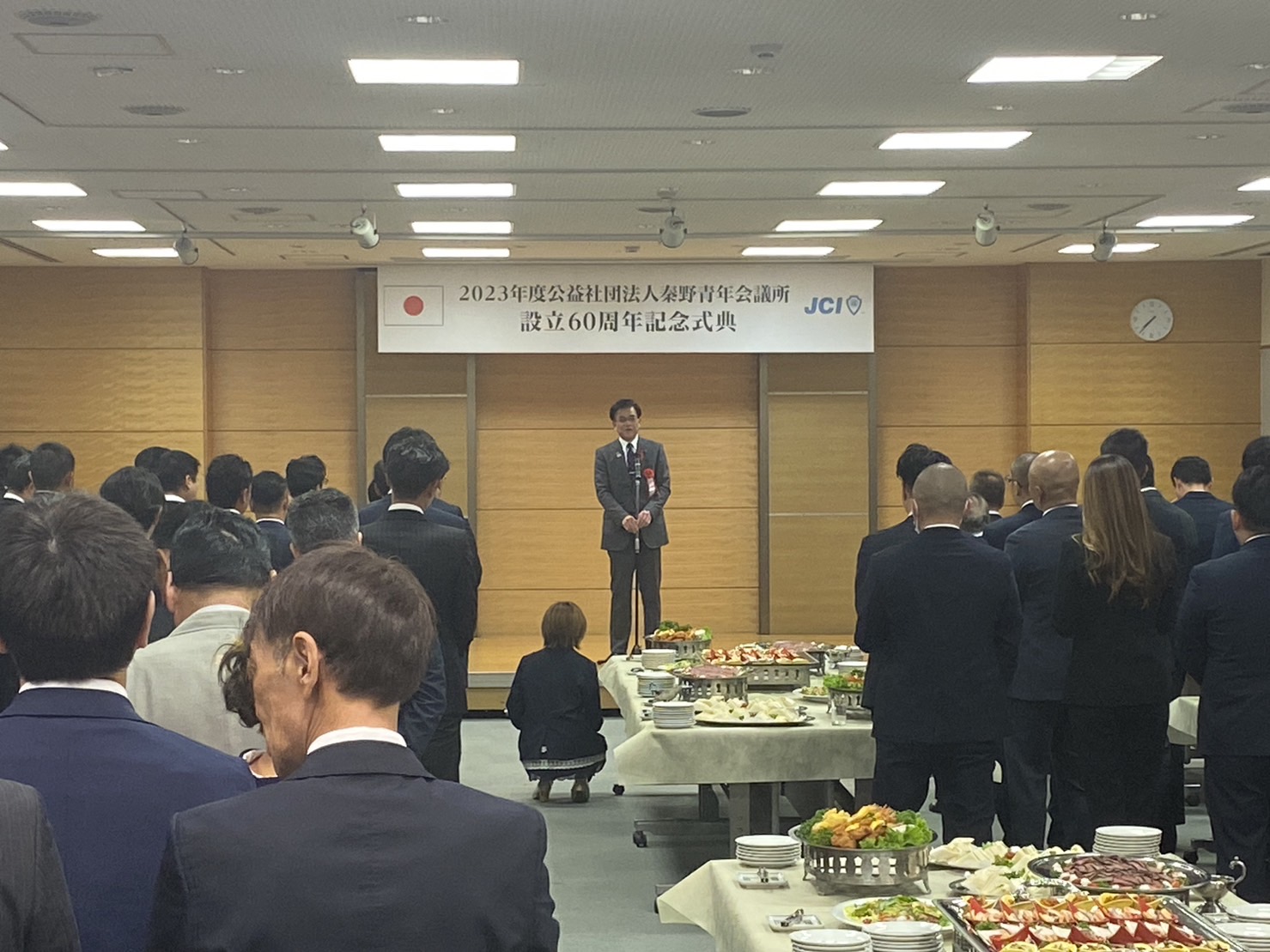 記念式典会場にて、市長が壇上で挨拶を行い、会場中央には料理が並び、周囲に多くのスーツ姿の参列者が立って話を聞いている式典の様子の写真