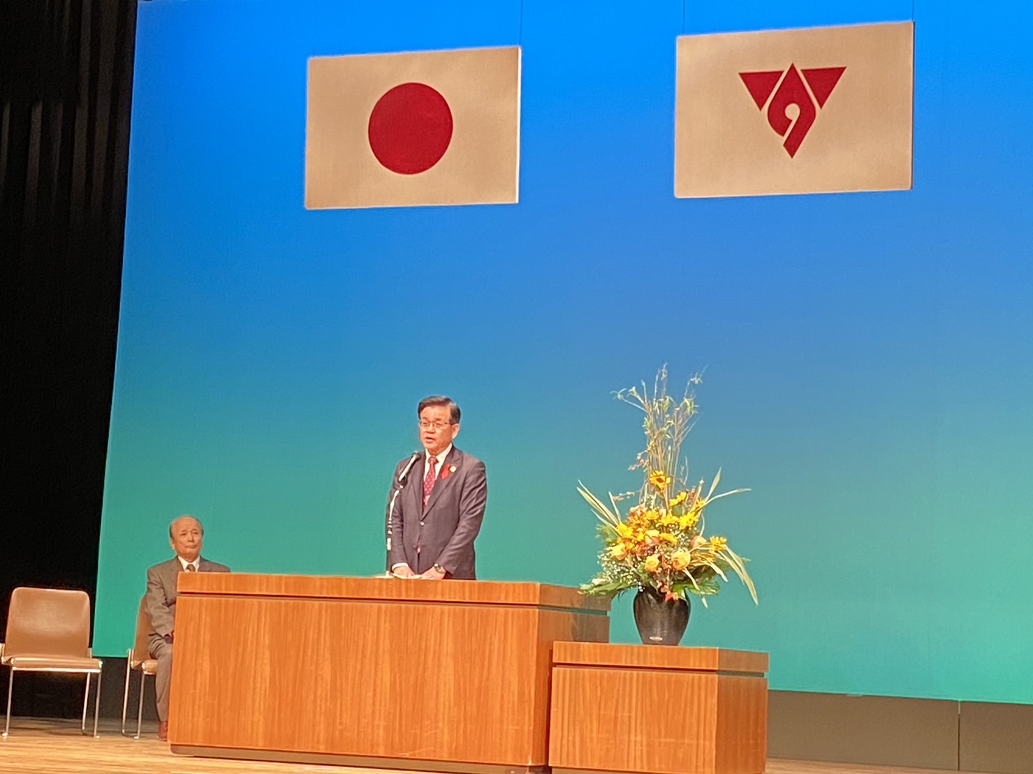 日本国旗と秦野市の市章が掲げられたステージで、きれいな生け花が飾られている演台の前に立つスーツ姿の市長がマイクの前で話をしている秦野市民会議の様子の写真