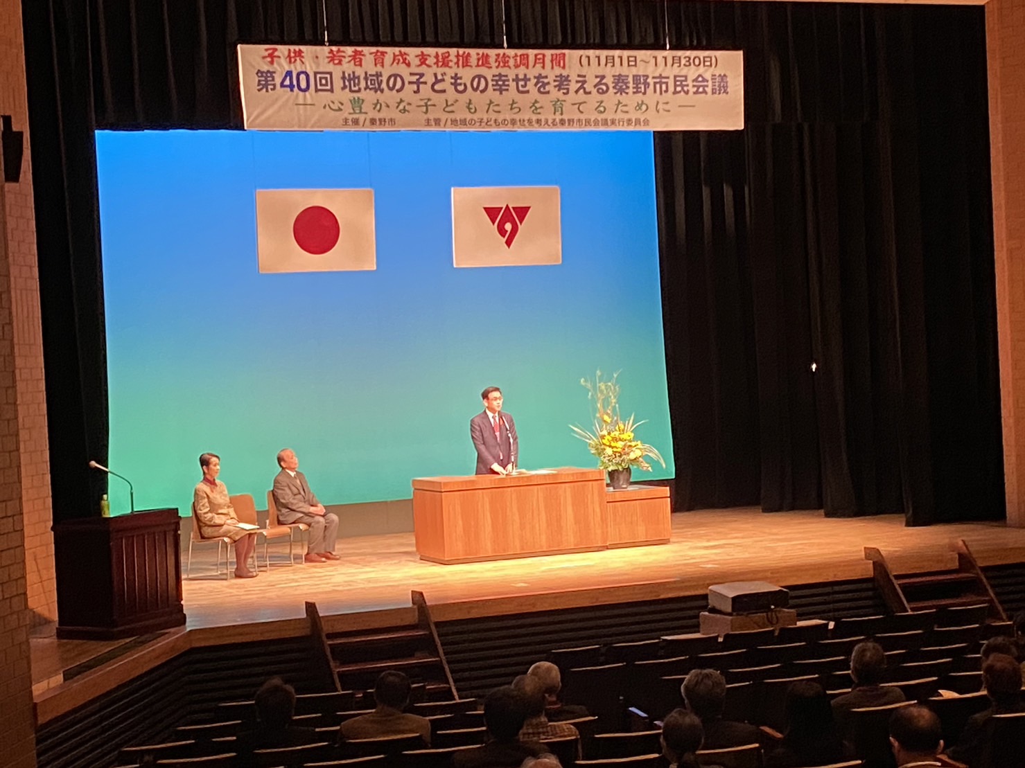 『第40回 地域の子どもの幸せを考える秦野市民会議』と書かれた横断幕が掲げられ、日本国旗と秦野市の市章が掲げられた舞台の中央に、きれいな生け花が飾られた演台があり、その前でスーツ姿の市長がマイクに向かって話をしており、後方には着席した2名の関係者が座り、客席には多くの市民が座っている会場全体の様子の写真