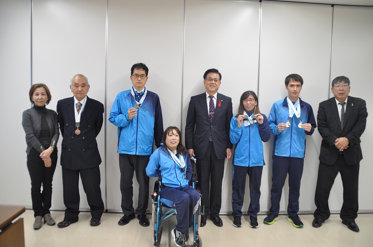 青いスポーツウェアを着た選手たちと、中央の車いすに座った選手、市長や関係者が並び、メダルを首にかけたり手に持って笑顔で写っている記念写真