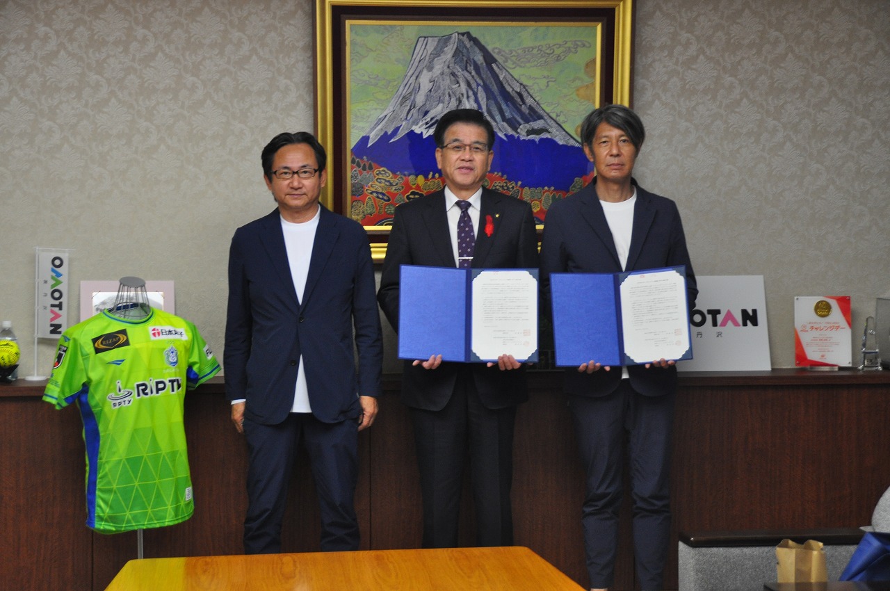 富士山の絵画を背景に、緑色のユニフォームがディスプレイされており、市長と右側の男性が締結書を持ち、左側に立つ関係者の男性と並んで写っている記念写真