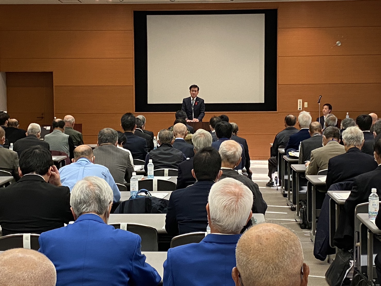 会議室の前方にある壇上で、黒いスーツを着た高橋市長がマイクの前で話しており、会場には多くの参加者が席に座り、話を聞いている様子を後ろから撮影した研修会の写真