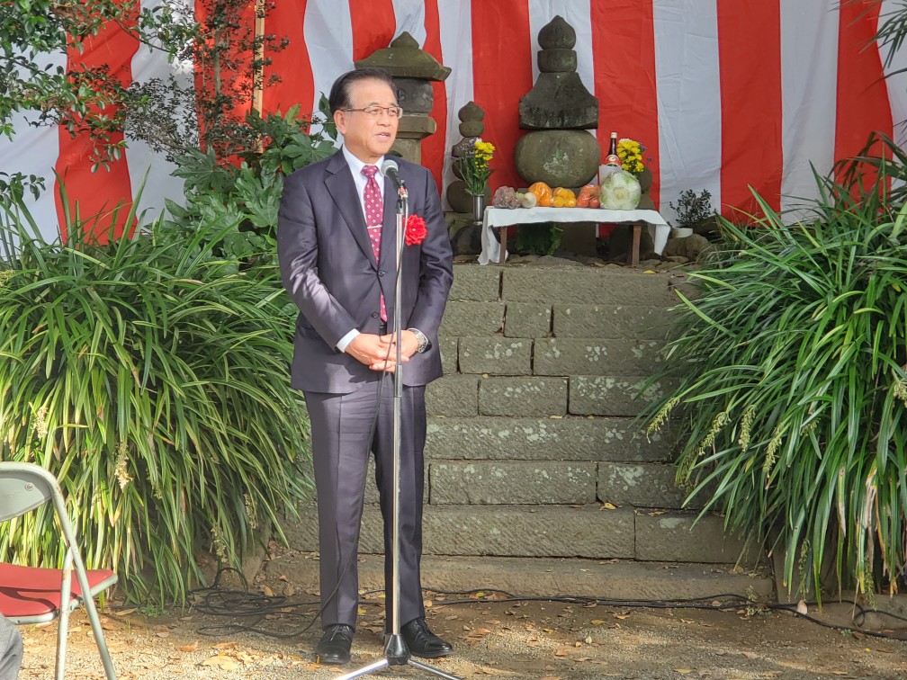 石段の上に供え物と石塔がある祭壇が設けられた屋外の式典会場で、紅白幕を背景にスーツ姿の市長がマイクの前に立ち挨拶をしている様子の写真