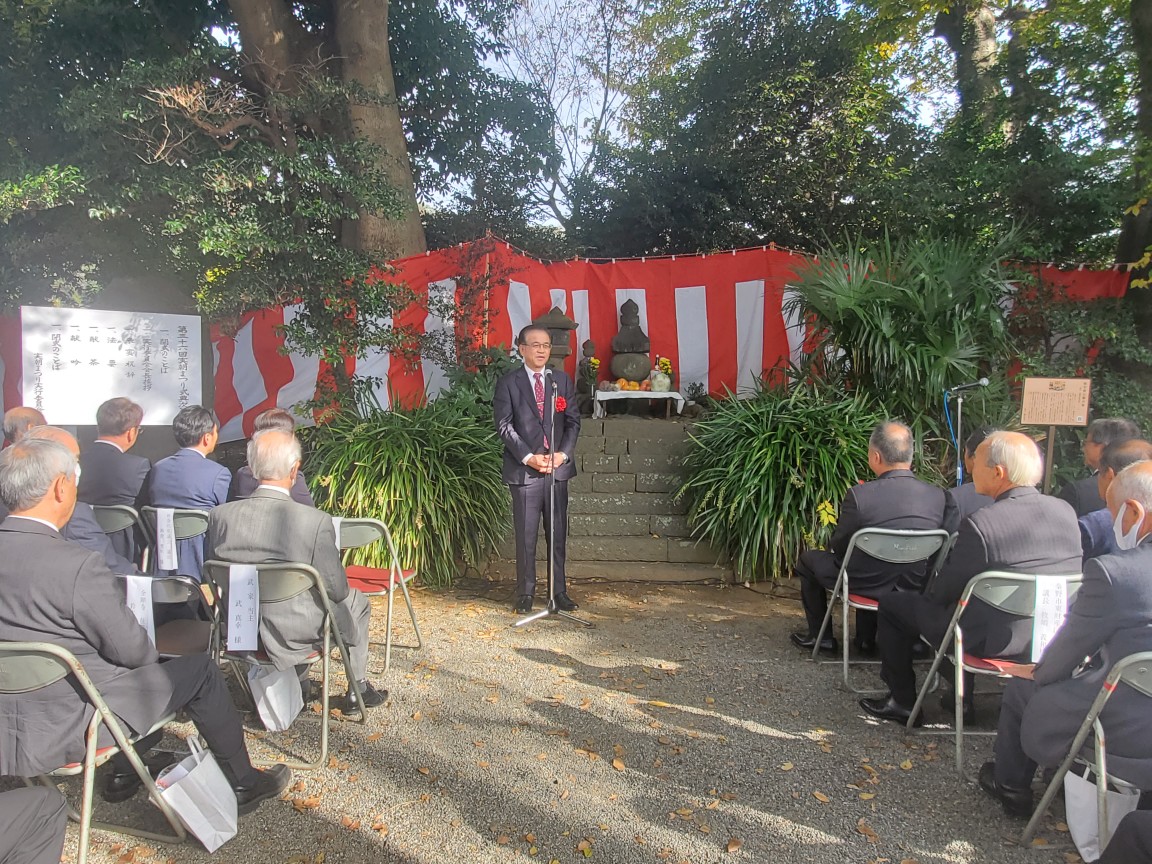 紅白幕と石塔を背景に設けられた屋外の祭壇前で、スーツ姿の市長がマイクの前で挨拶をしており、その前に椅子に座った多くの参列者たちが話を聞いている式典の様子の写真