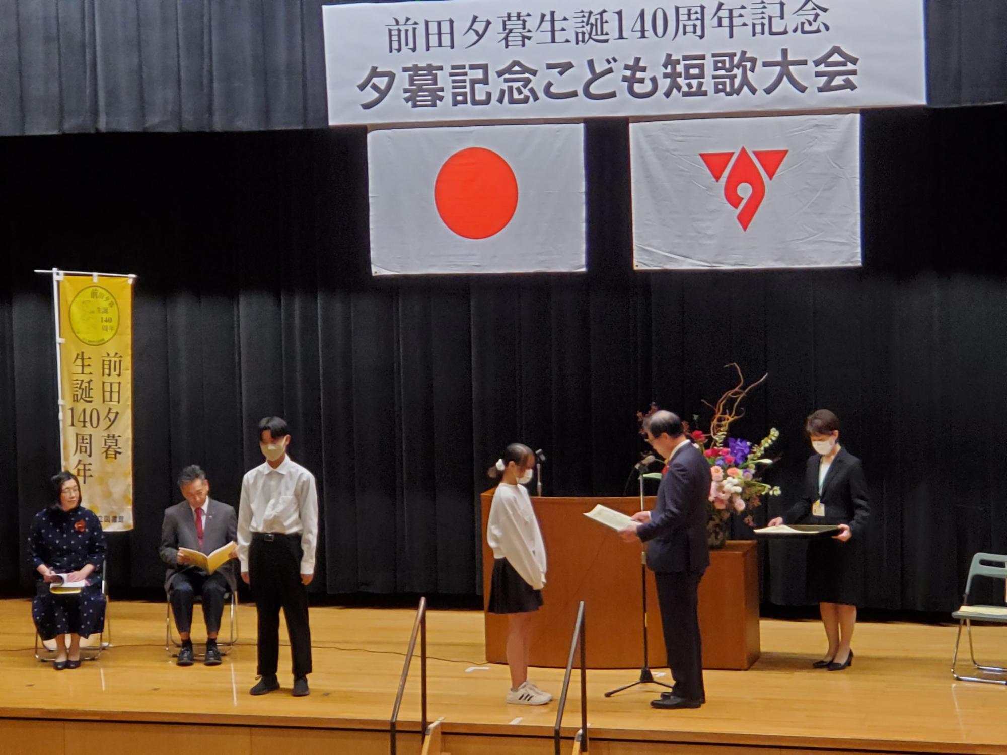 『前田夕暮生誕140周年記念夕暮記念こども短歌大会』書かれた横断幕が掲げられ、日本国旗と秦野市の市章が掲げられた舞台で、受賞者の女子生徒が市長から表彰を受けている様子を写した写真