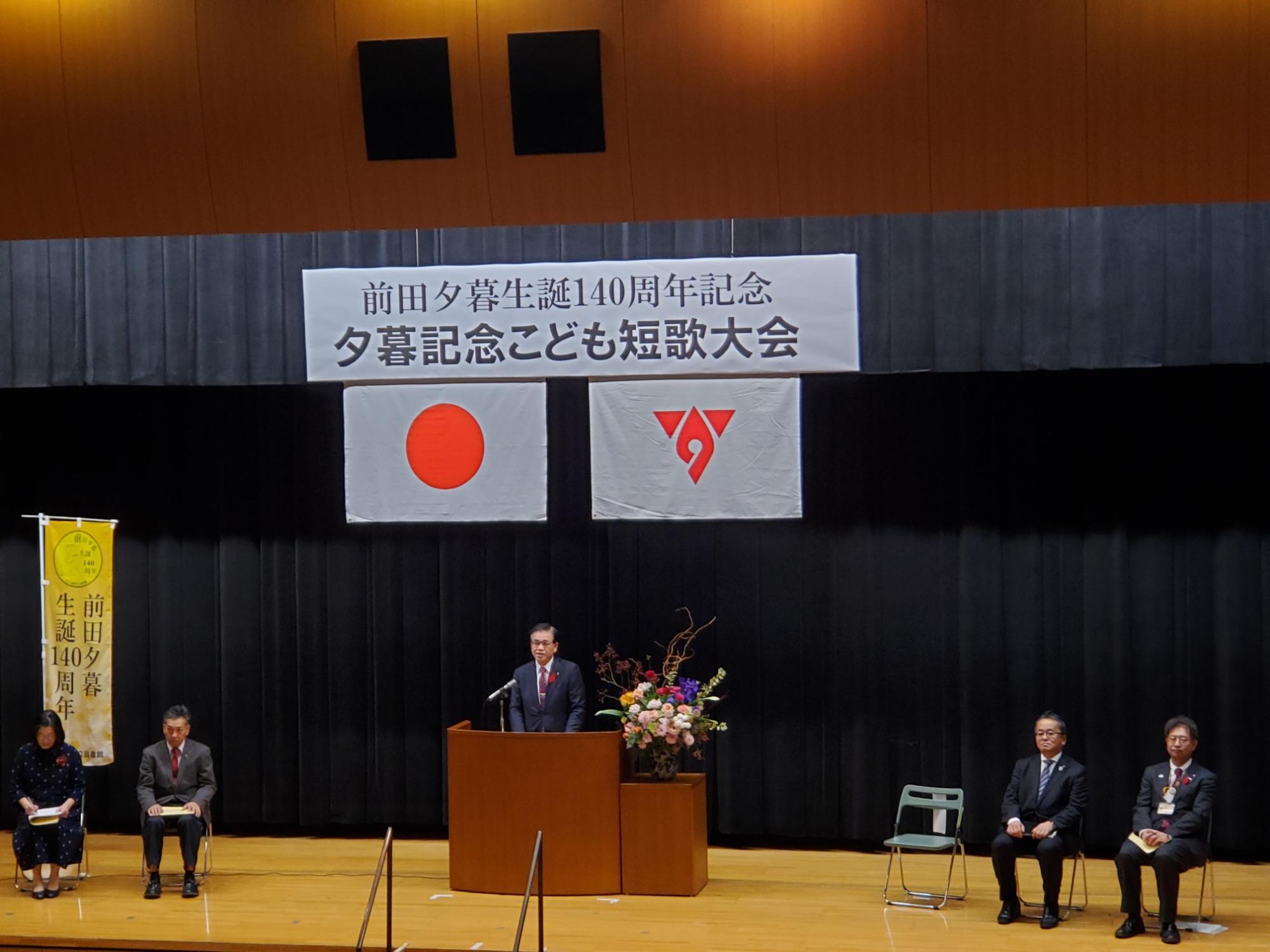 『前田夕暮生誕140周年記念夕暮記念こども短歌大会』書かれた横断幕が掲げられ、日本国旗と秦野市の市章が掲げられた舞台で、関係者の方が舞台上の椅子に数名座っており、中央の檀上で市長が話をしている表彰式の様子の写真