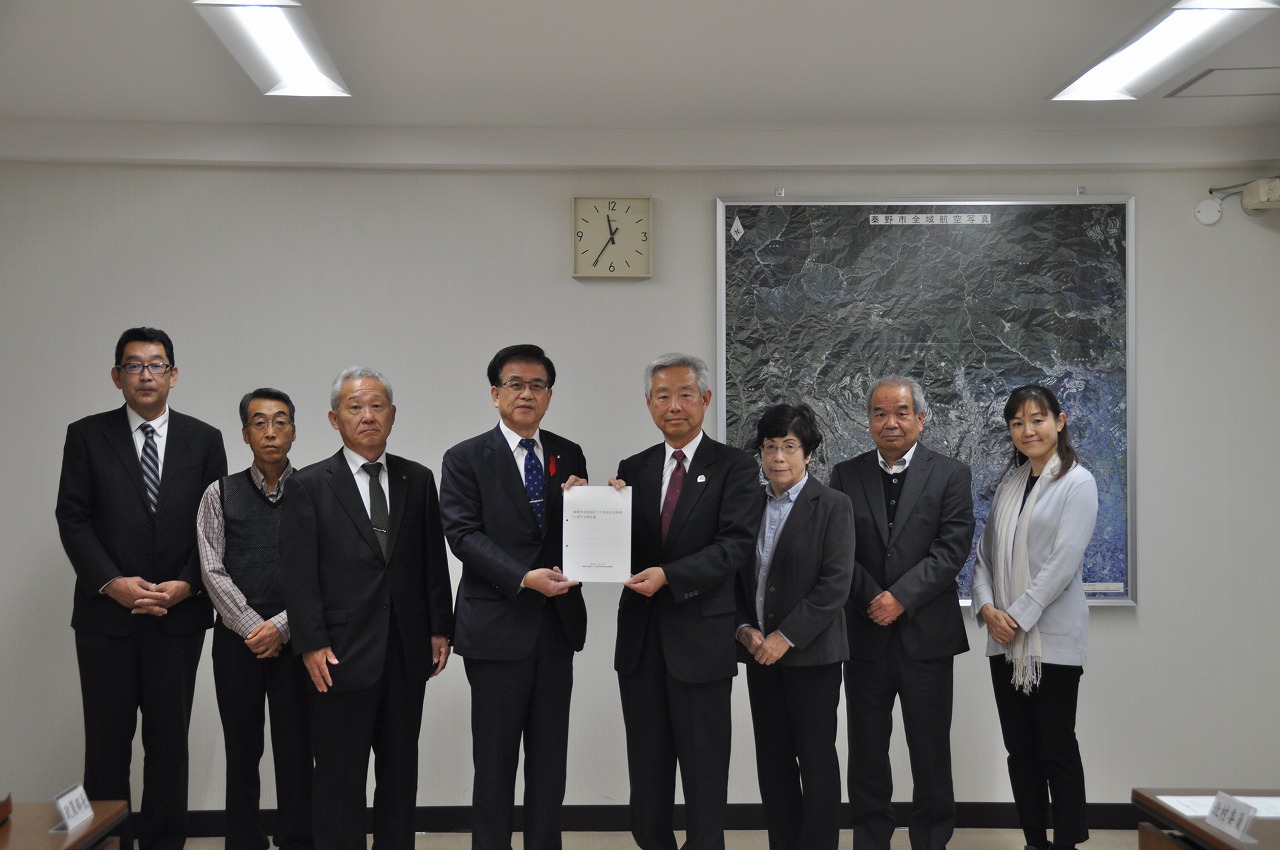 中央に立つ市長と関係者の男性が報告書を一緒に持ち、委員の方々と横一列に並び、写っている集合写真