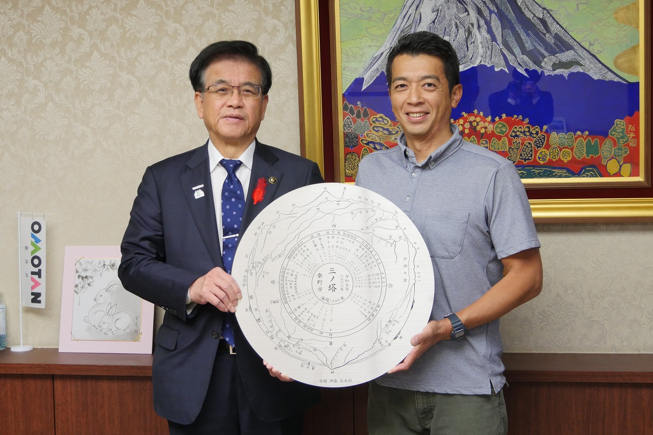 富士山の絵画を背景に、市長と伊藤代表取締役が三ノ塔山座同定盤を一緒に持ち、笑顔で写っている記念写真