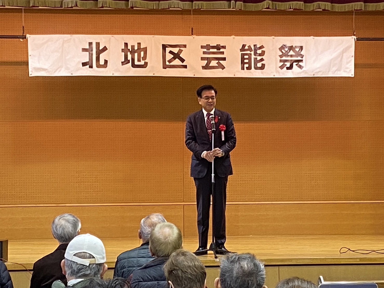 『北地区芸能祭』の横断幕が掲げられた会場の舞台で、スーツ姿の市長がマイクの前に立ち、観客に向かって話している様子の写真