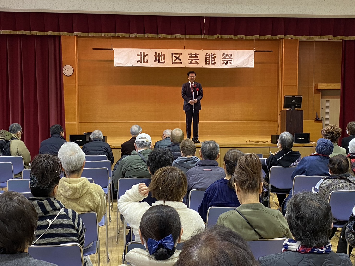北地区芸能祭の横断幕が掲げられた会場の舞台で、スーツ姿の市長がマイクの前に立ち話をしており、客席には多くの来場者が席について話をしている様子を後方から写した写真
