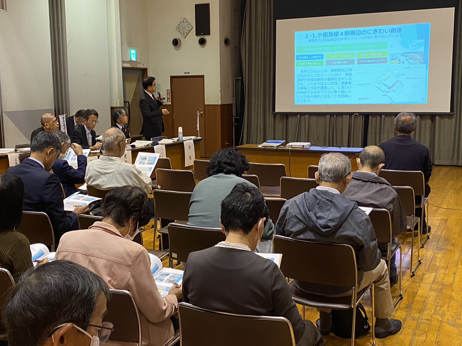 前方のスクリーンに資料を映しだし、市長がマイクを持って説明しており、席についている参加者の方々が資料に目を通しながら話を聞いている様子の写真