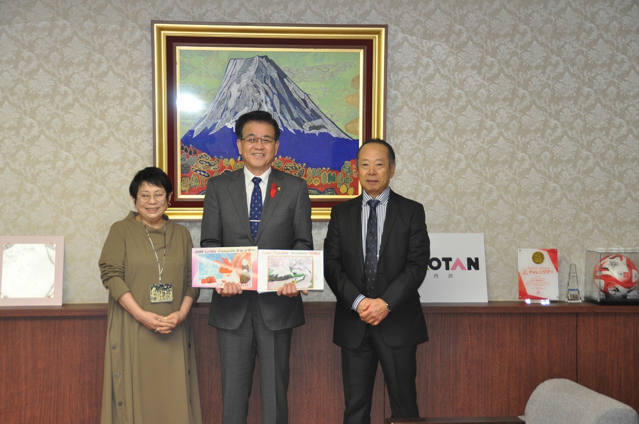 富士山の絵画を背景に、寄贈された絵本を2冊持った市長、久保寺さん、関係者の男性が並び、笑顔で写っている記念写真