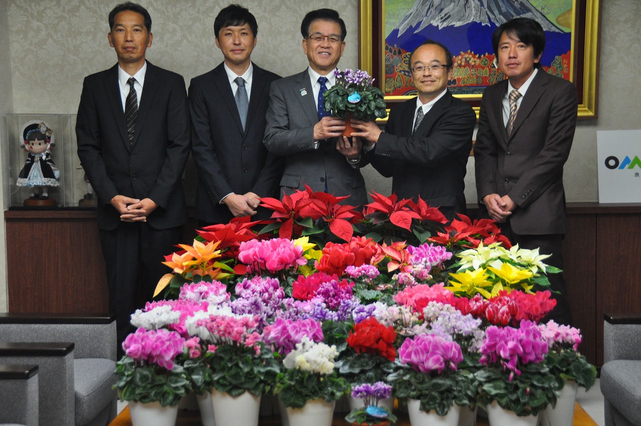 富士山の絵画を背景に、贈されたピンクや白、紫色など鮮やかな色彩のシクラメンの鉢植えを前に、JAはだの花き部会鉢物部の相原部長と市長、関係者の方々5名が並んで写っている記念写真