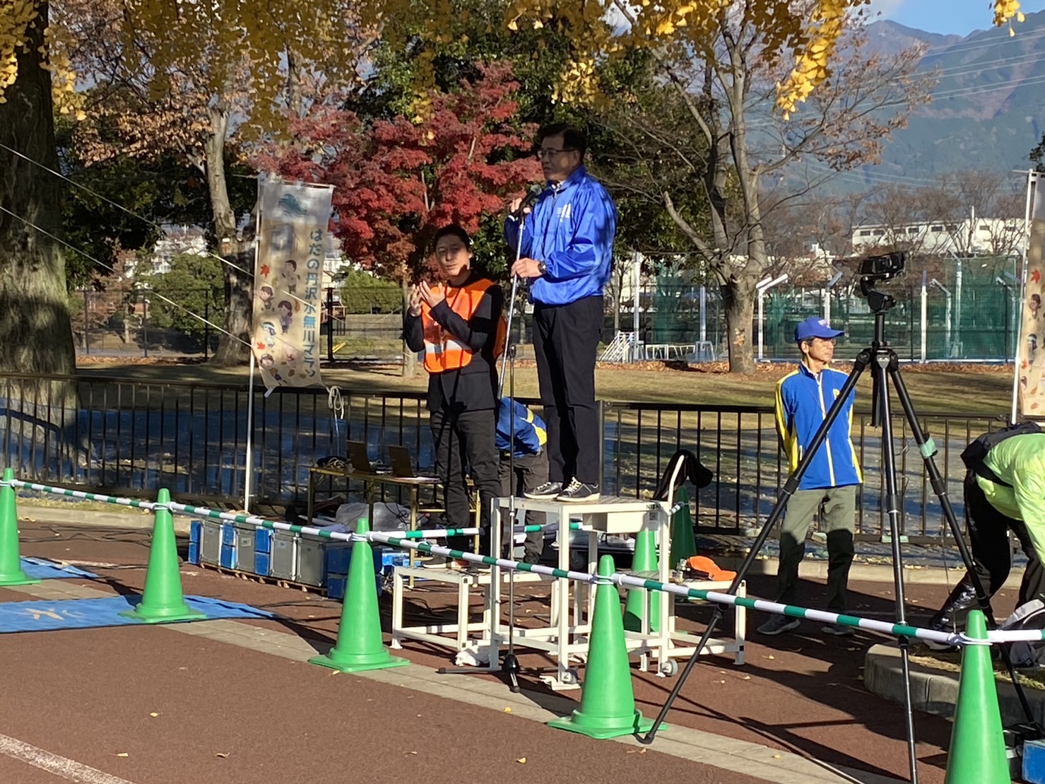 秋の紅葉に包まれたマラソン大会会場で、青いジャンパーを着た市長が台の上に立ち、マイクの前で話をしている様子の写真
