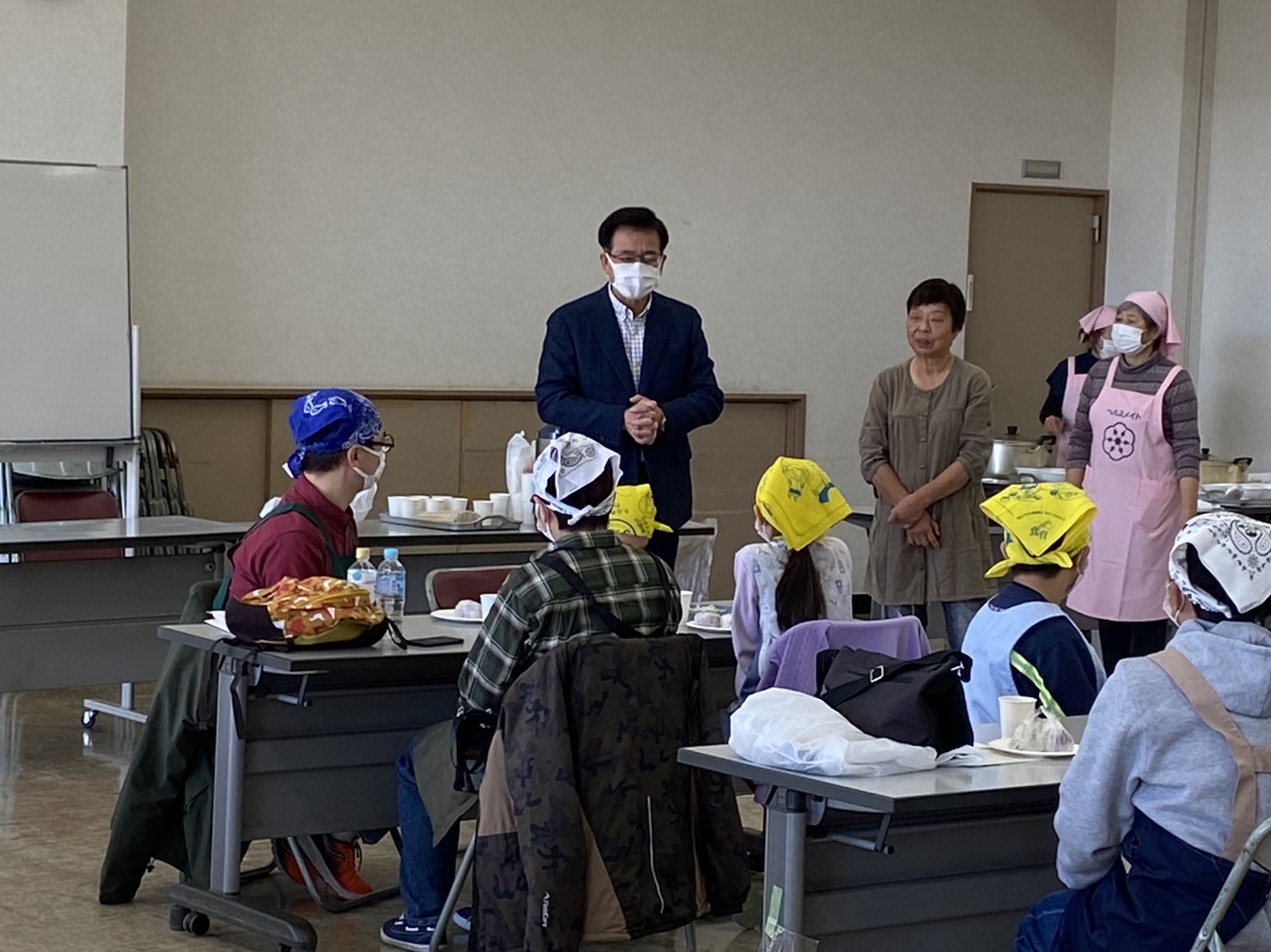 頭にバンダナを巻き、マスクとエプロンを身に着けた参加者親子が席についており、マスク姿の市長が参加者の皆さんに話をしている様子の写真