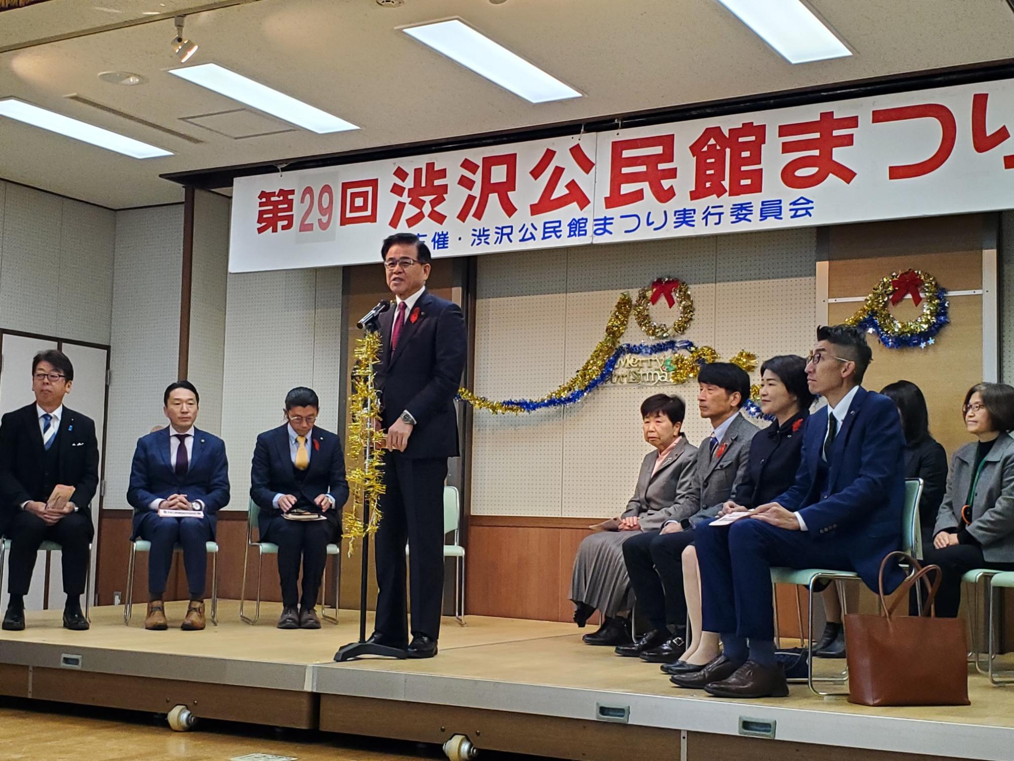 「第29回 渋沢公民館まつり」と書かれた横断幕が掲げられた会場の舞台上には正装をした来賓の方々椅子に座っており、スーツ姿の市長が金色のモールで飾られたマイクの前に立って話をしている渋沢公民館まつりの様子の写真