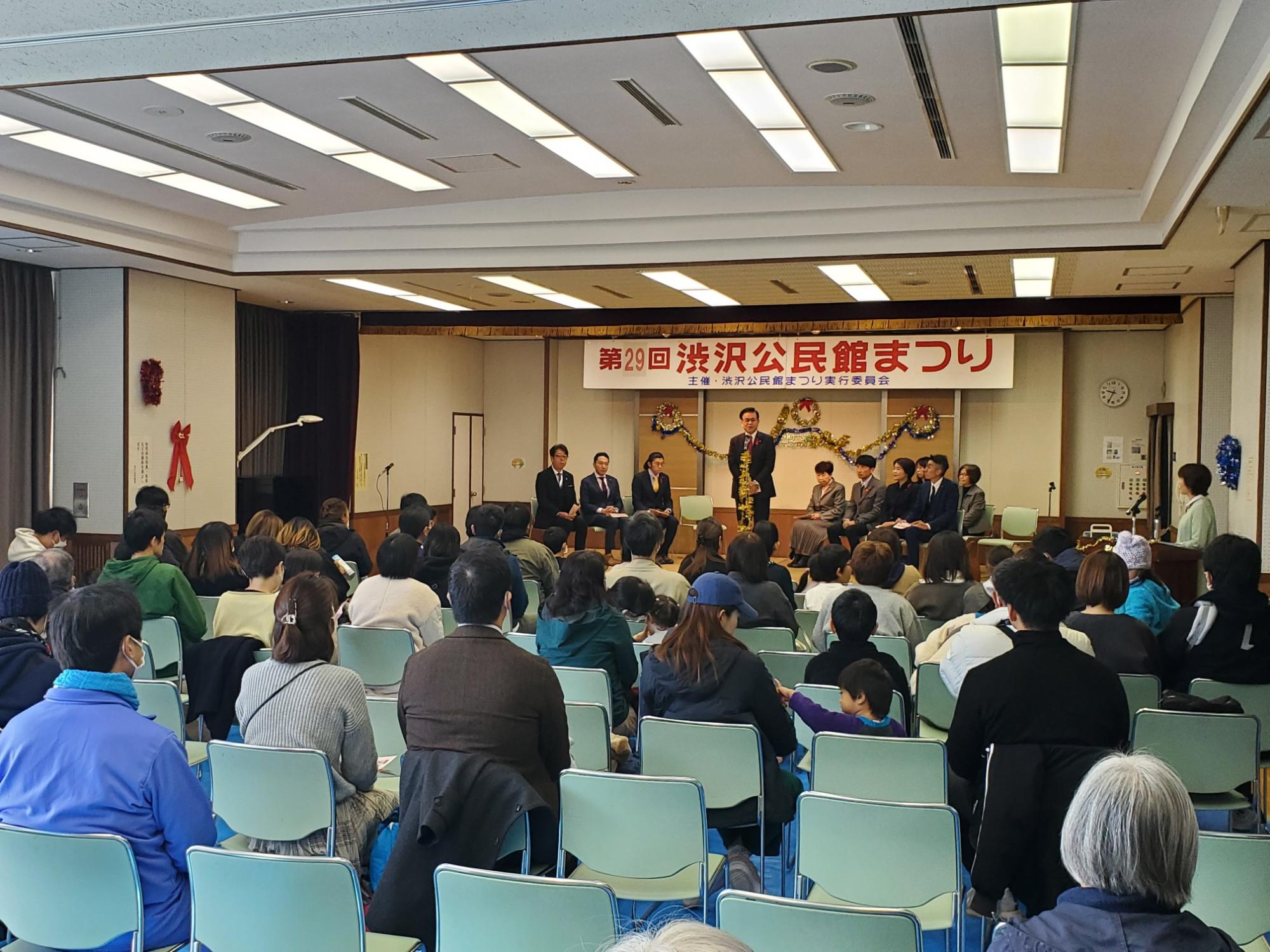 「第29回 渋沢公民館まつり」の横断幕が掲げられた会場の舞台で話をしている市長と、多くの来場者が席についている様子を後方から撮影した会場全体の写真
