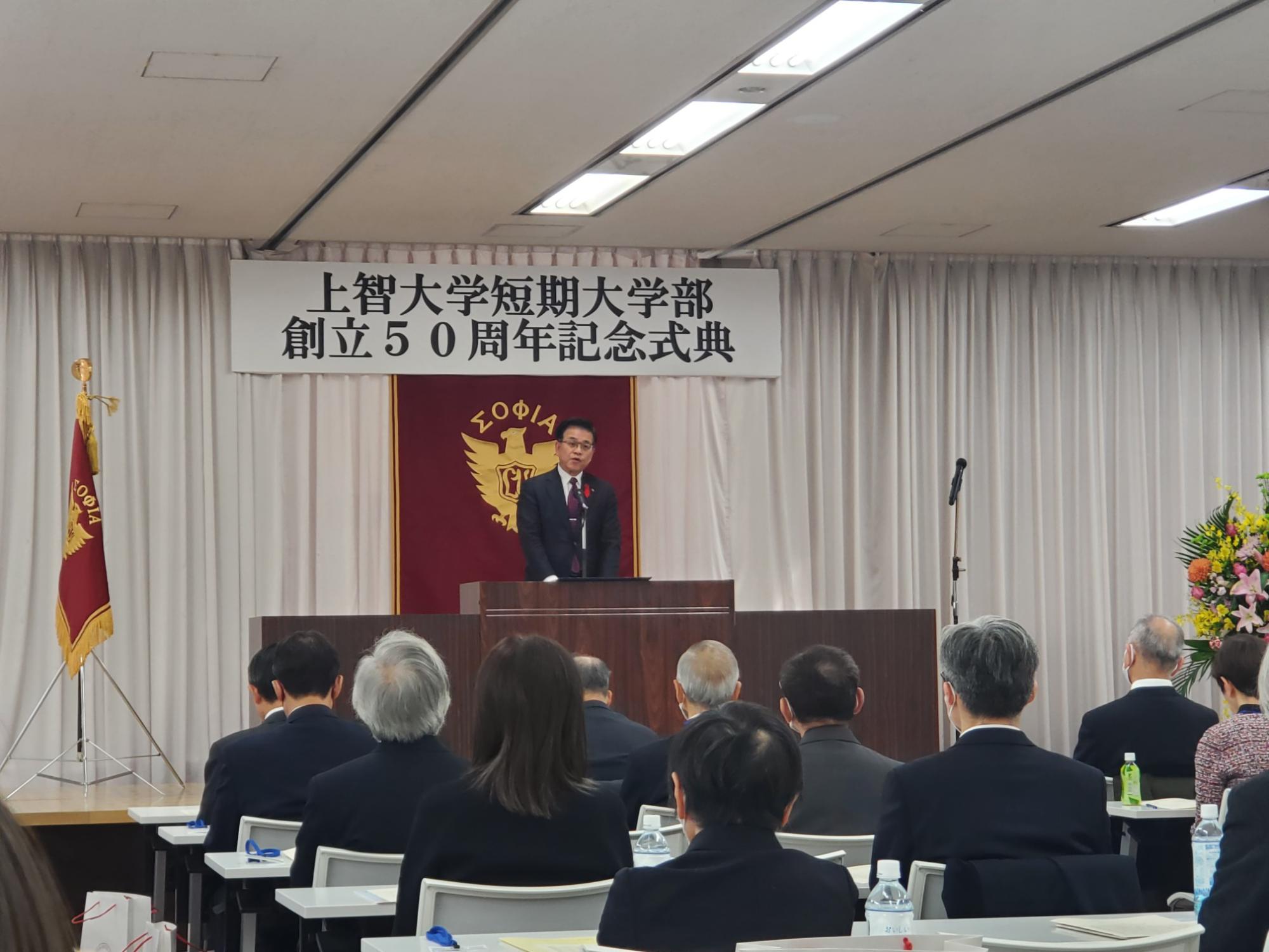 「上智大学短期大学部 創立50周年記念式典」と書かれた横断幕と、赤地に金色の校章が掲げられた舞台の壇上で、市長がスピーチをしており、会場にはスーツ姿の多くの参加者が着席して話を聞いている式典の様子を写した写真