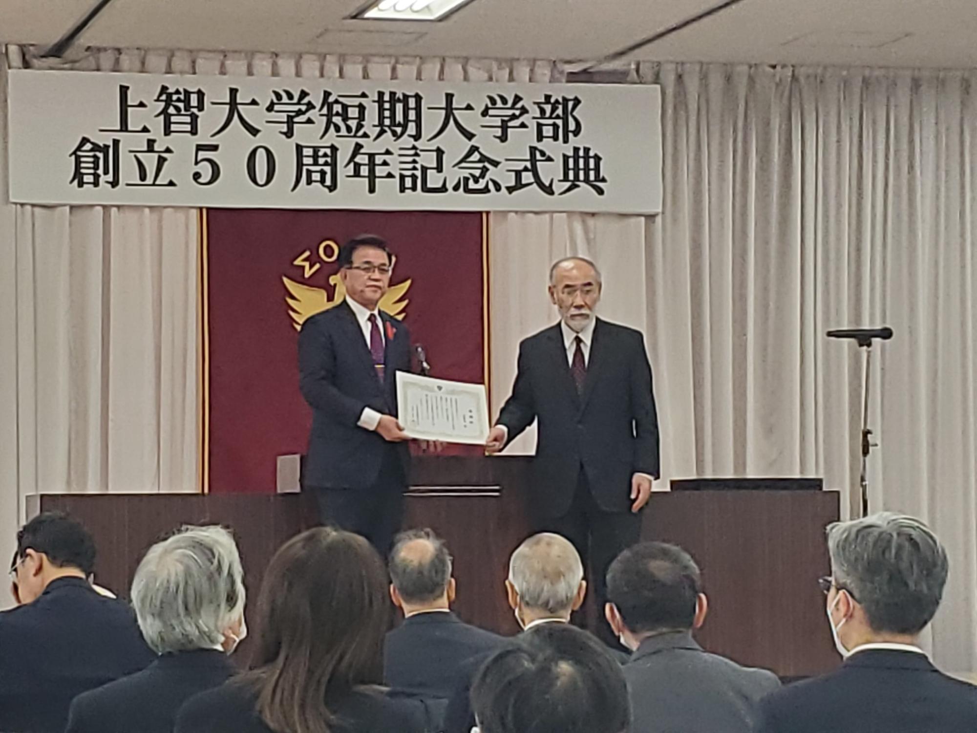 「上智大学短期大学部 創立50周年記念式典」と書かれた横断幕が掲げられた壇上で、赤地に金色の校章を背景に、スーツ姿の市長と関係者の男性が賞状を一緒に持って並んでおり、会場にはスーツ姿の多くの参加者が着席している式典の様子を写した写真