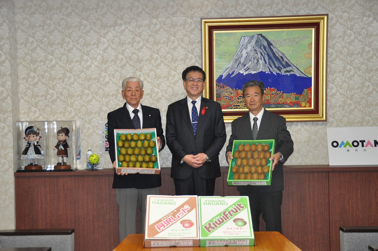 富士山の絵を背景に笑顔の高橋市長と両側には贈答用のキウイフルーツの箱を持った石原部長と関係者の男性が並んで写っている記念写真